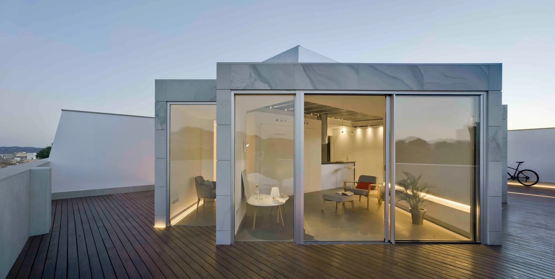 A modern rooftop terrace features dark wood decking and a gray stone-clad structure with floor-to-ceiling glass sliding doors showing a minimalist interior.