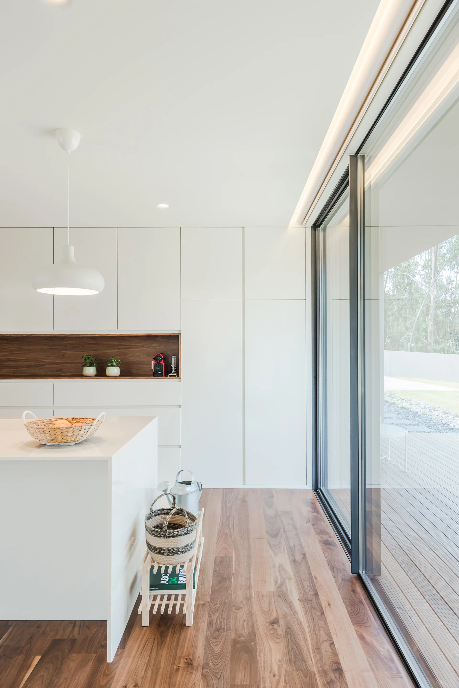 Minimalist white kitchen featuring seamless cabinetry with a wood accent niche, a white island, and large sliding glass doors leading to an outdoor deck.