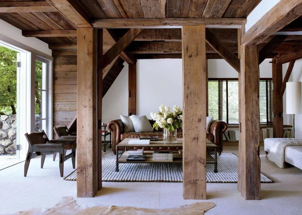 A rustic living room viewed through heavy vertical wood timber columns, featuring a tufted brown leather sofa, wood coffee table, and light stone flooring.