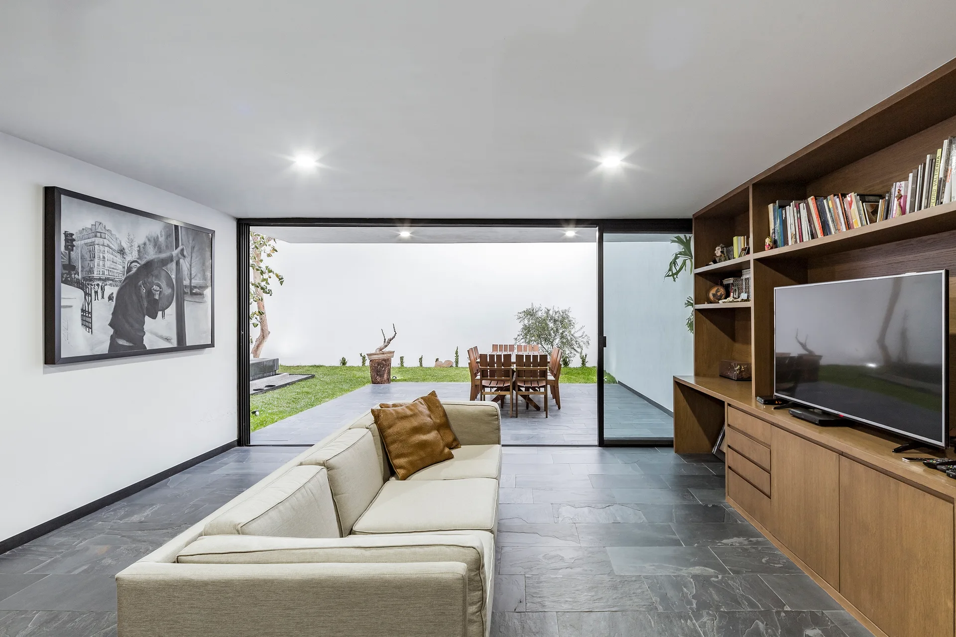 A modern living room featuring a cream sofa, a large wood built-in shelving unit with a TV, and slate tile floors that extend to an outdoor patio.