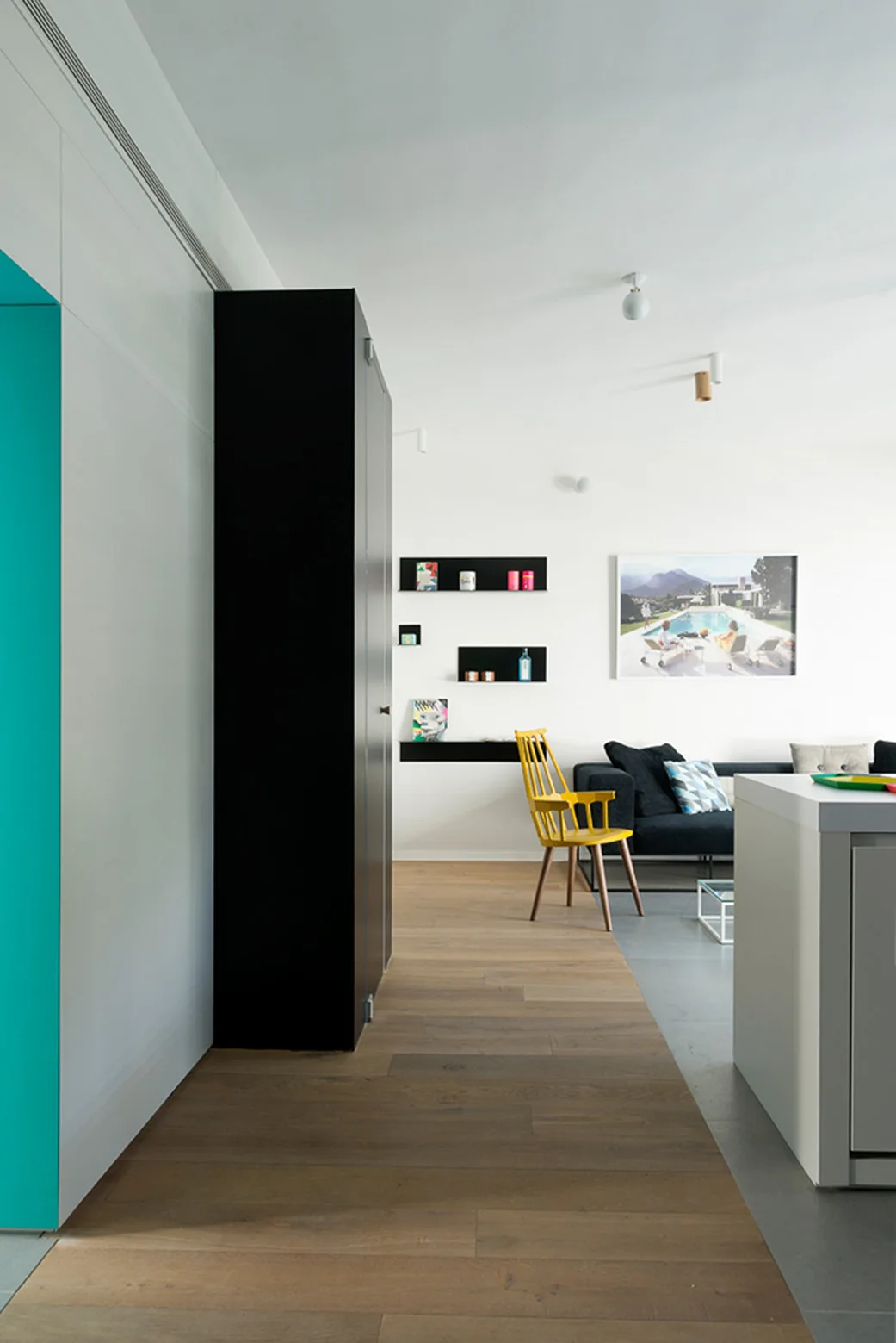 A modern living room features a dark sofa, a bright yellow wooden chair, black floating shelves, and a tall black cabinet on wood flooring.