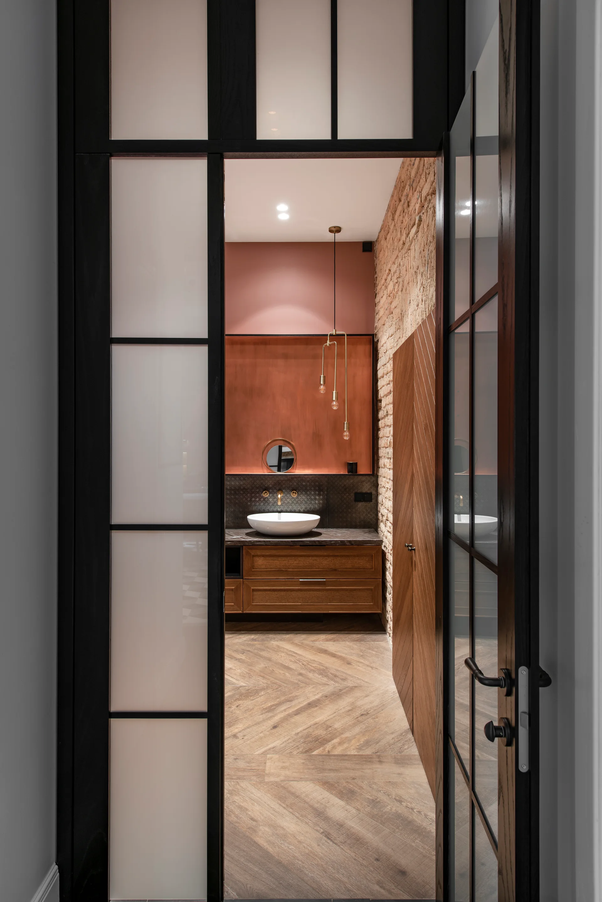A view through a black-framed glass door into a bathroom with a wood vanity, white vessel sink, herringbone flooring, and an exposed brick wall.