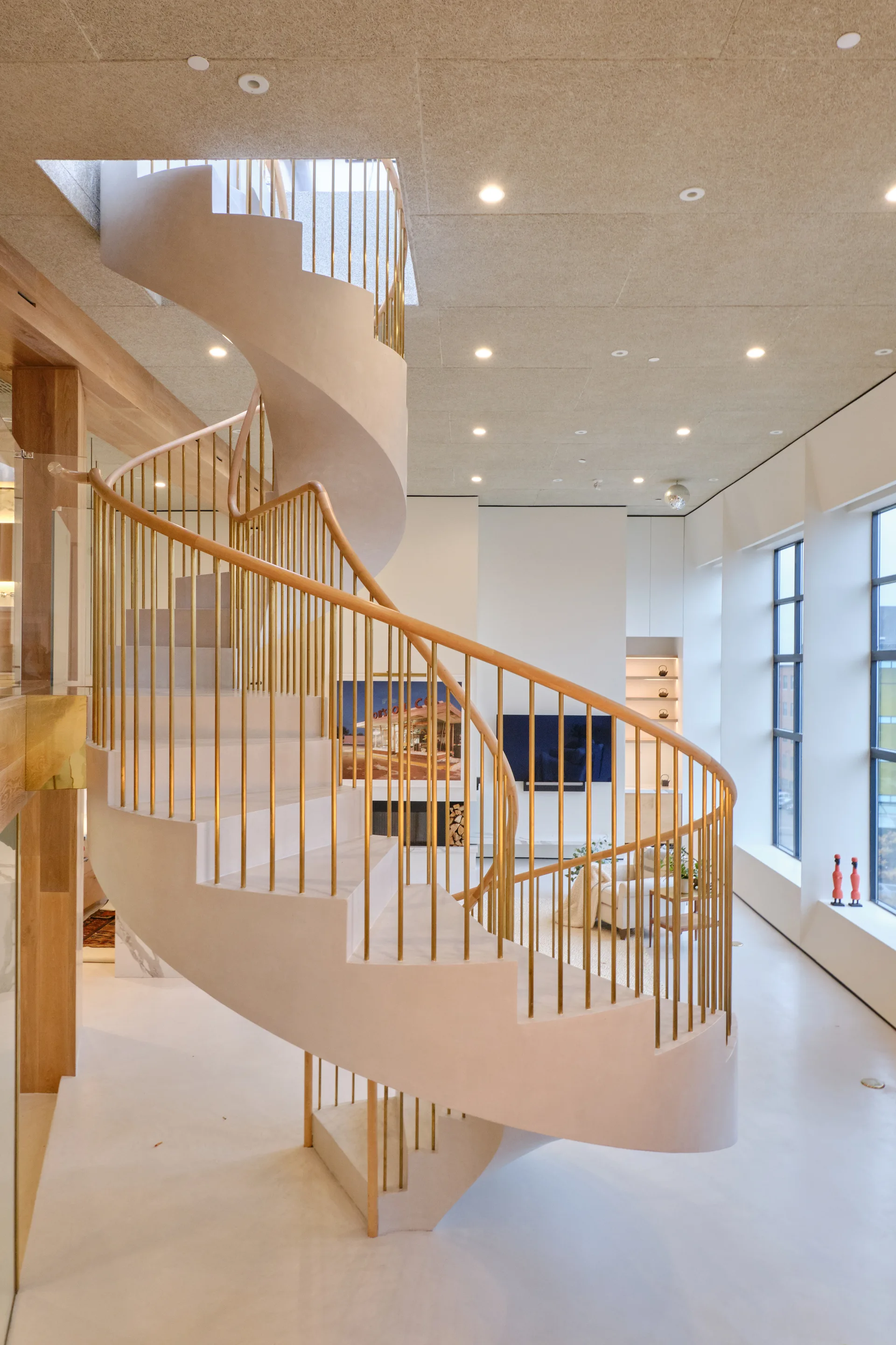 A sculptural beige plaster spiral staircase with vertical brass balusters and a wood handrail stands in an open modern living room.