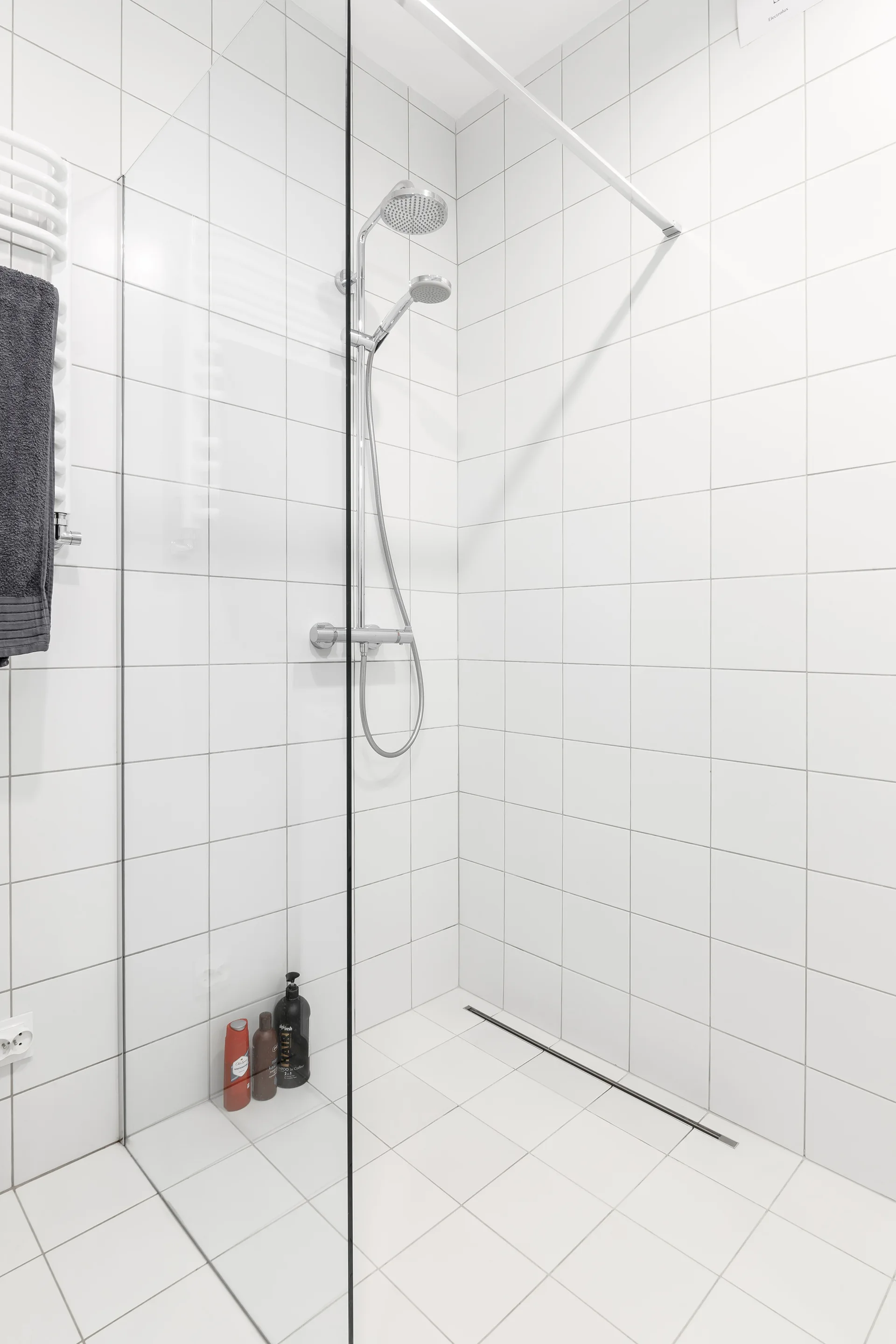 A minimalist walk-in shower features white square wall tiles, a glass partition, chrome fixtures, and a linear floor drain.