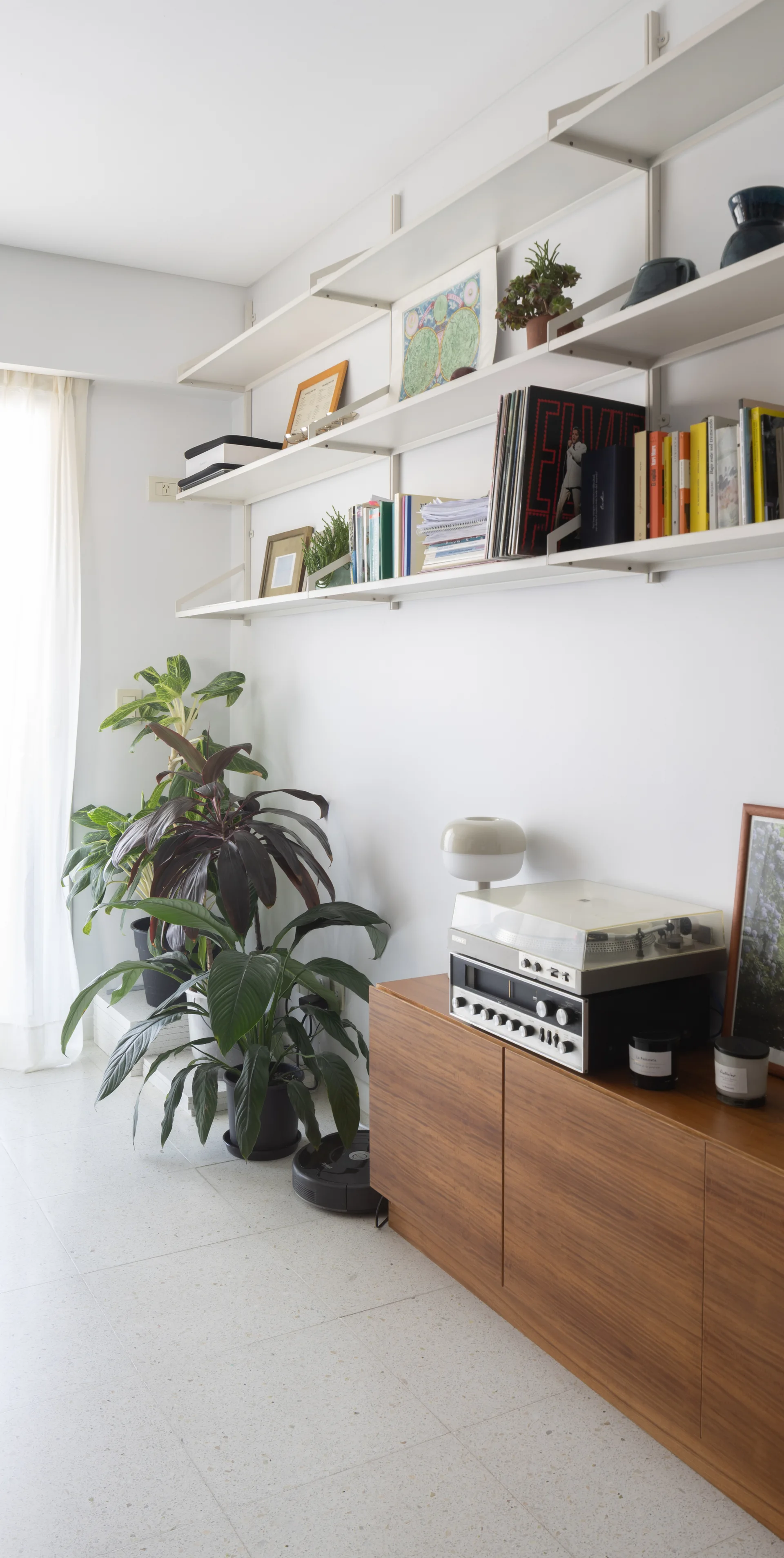 A wooden console table with a vintage record player stands on terrazzo flooring beneath long white wall shelves filled with books and albums.