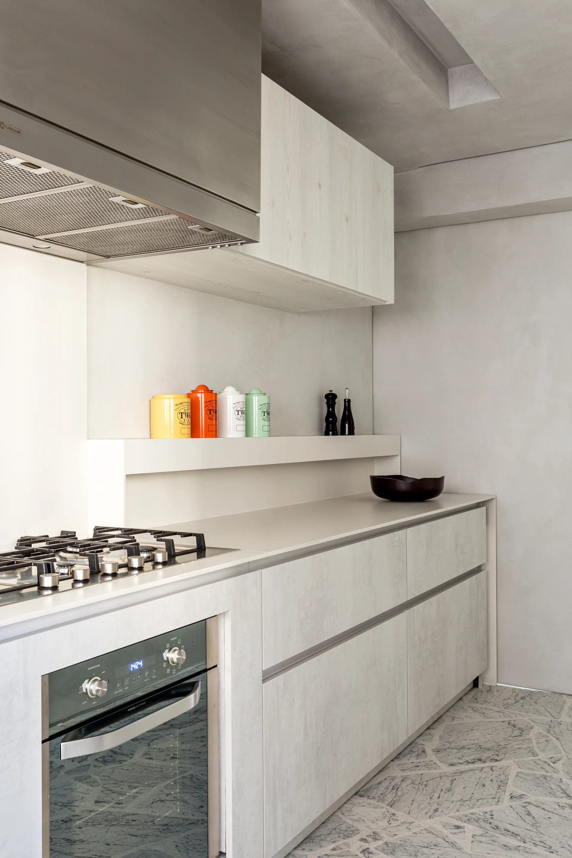 A modern kitchen features light gray lower cabinets, a stainless steel range hood, a built-in oven, and colorful canisters on a recessed ledge.