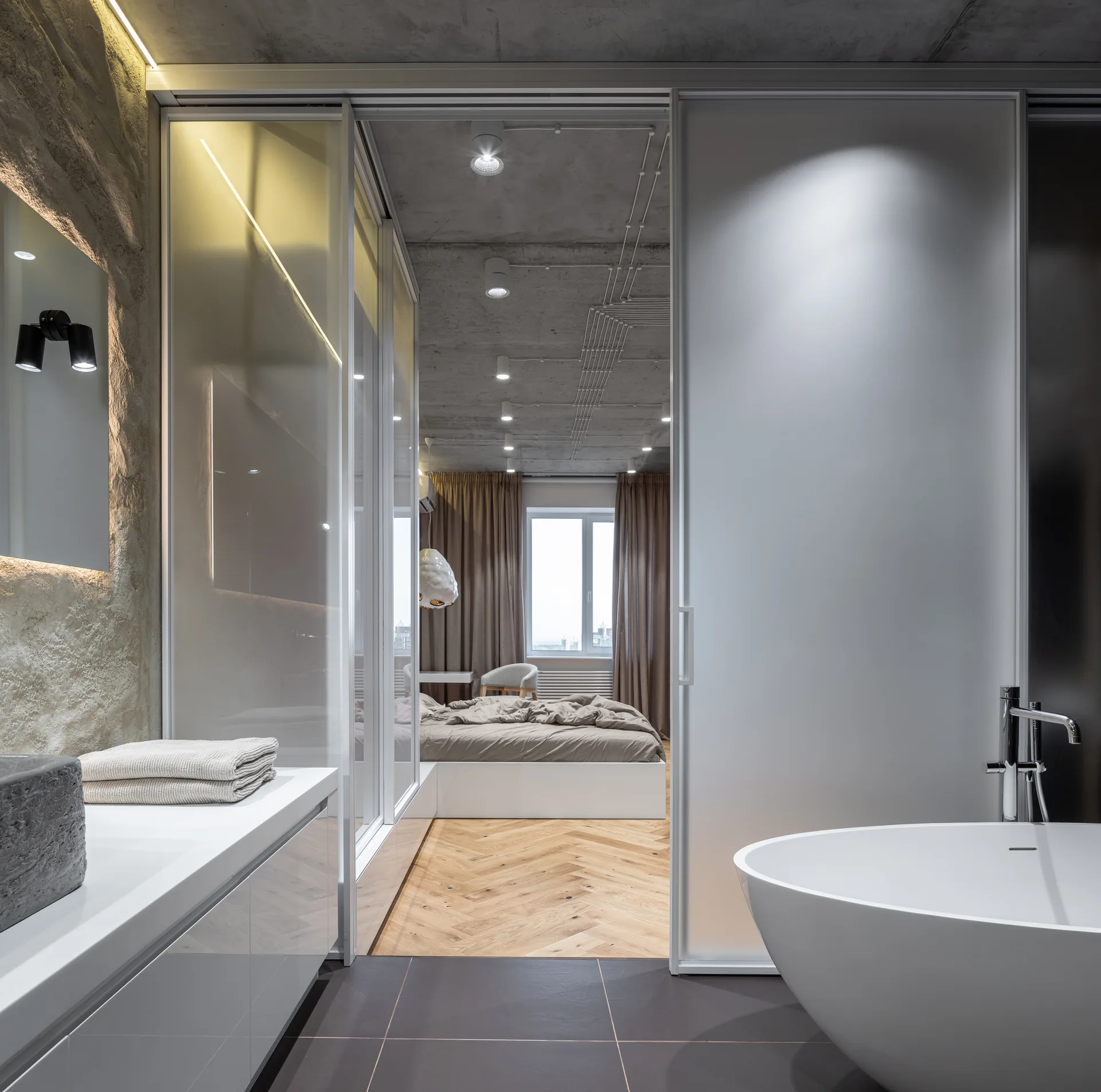 A modern bathroom featuring a white freestanding tub views an adjacent bedroom through a frosted sliding glass door.