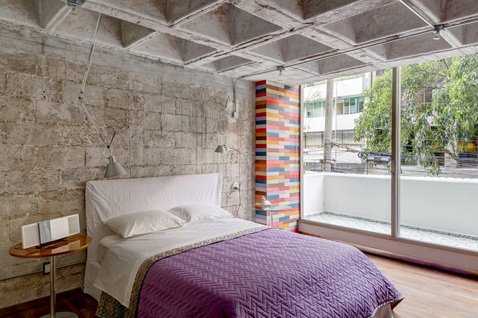 An industrial bedroom features an exposed concrete waffle ceiling, a block wall, a multi-colored tiled column, and a bed with a purple quilt.