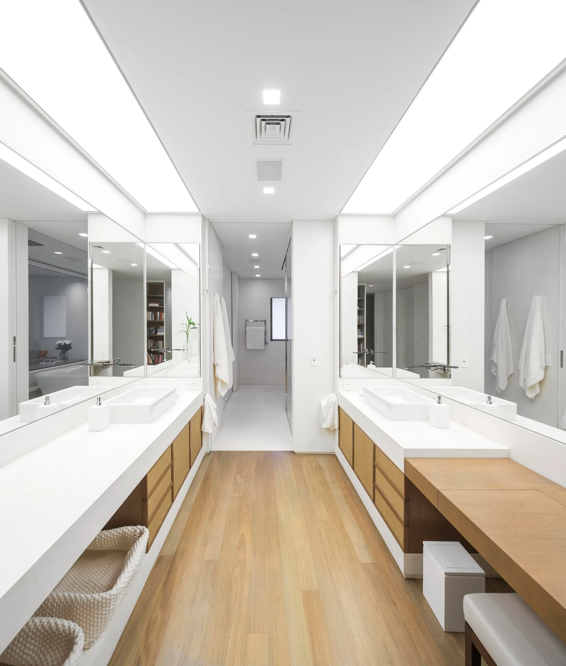 A symmetrical modern bathroom featuring dual white vanities with wood cabinets, large mirrors, wood flooring, and bright overhead coved lighting.