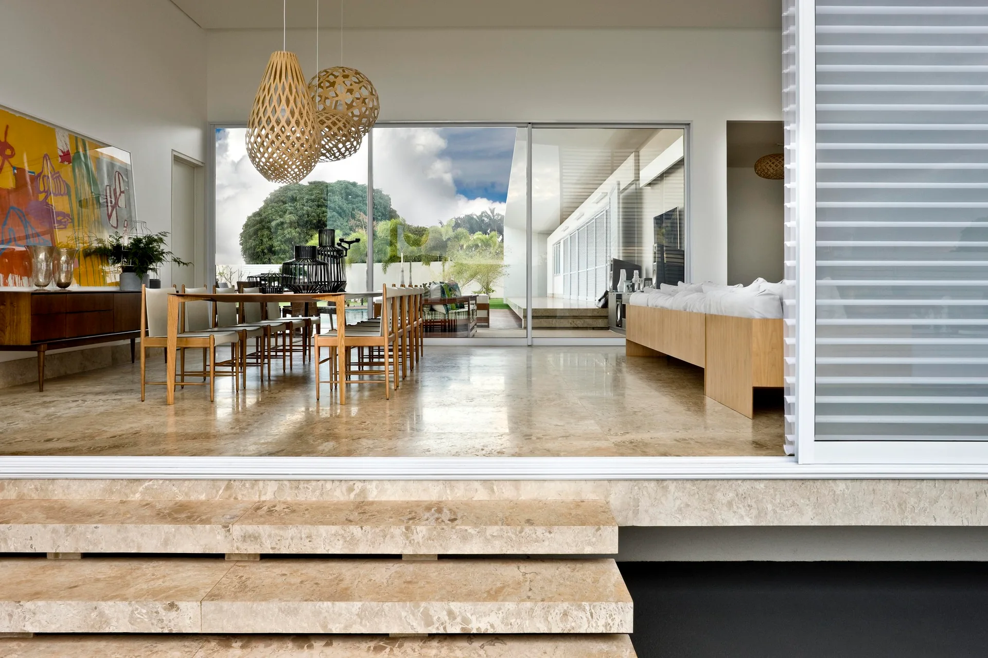 A spacious modern dining room features a wooden table and chairs, large woven pendant lights, polished stone flooring, and sliding glass doors.