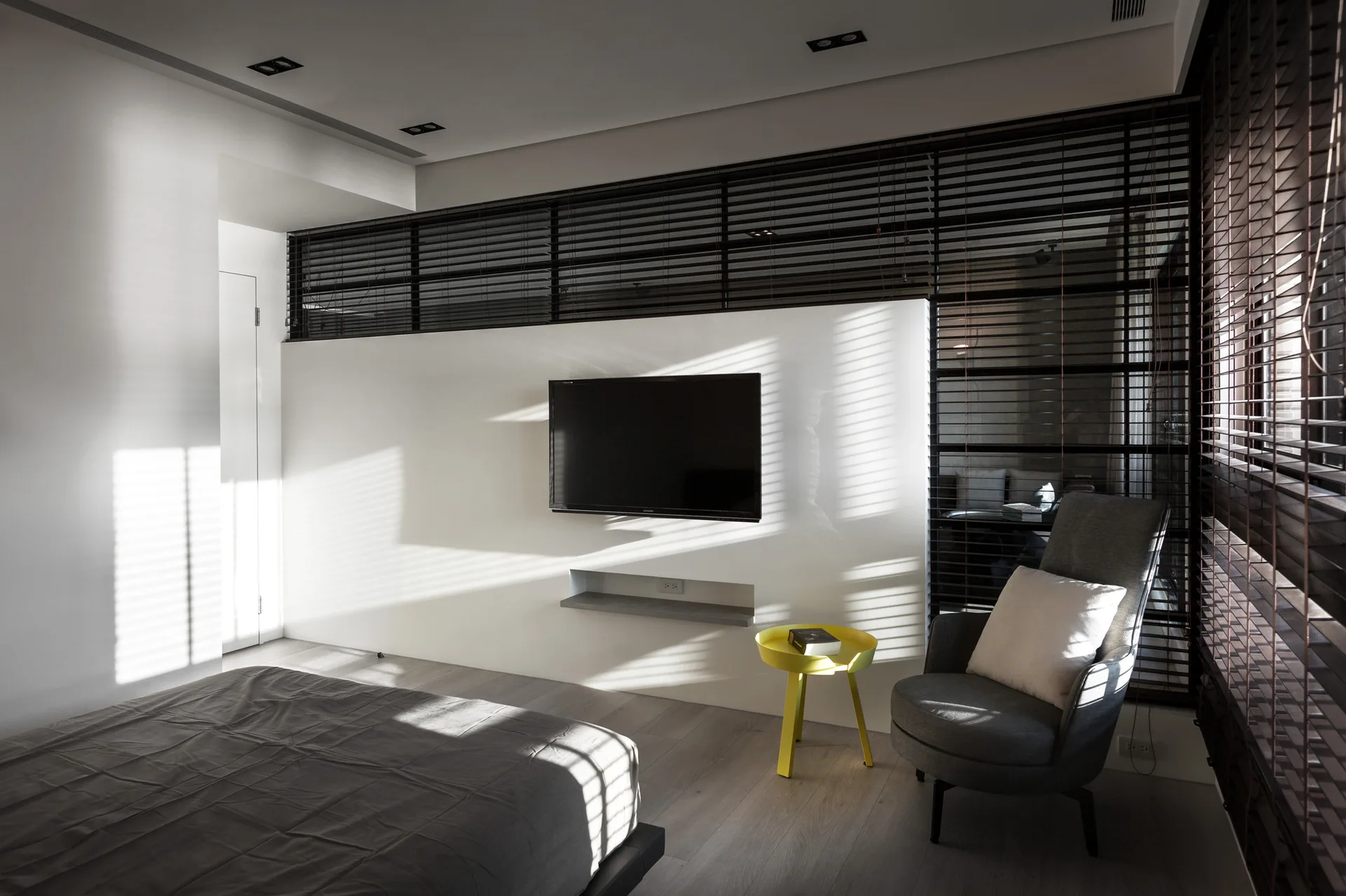 A modern minimalist bedroom featuring a grey bed, a white partition wall with a mounted TV, a grey armchair, and a bright yellow side table.