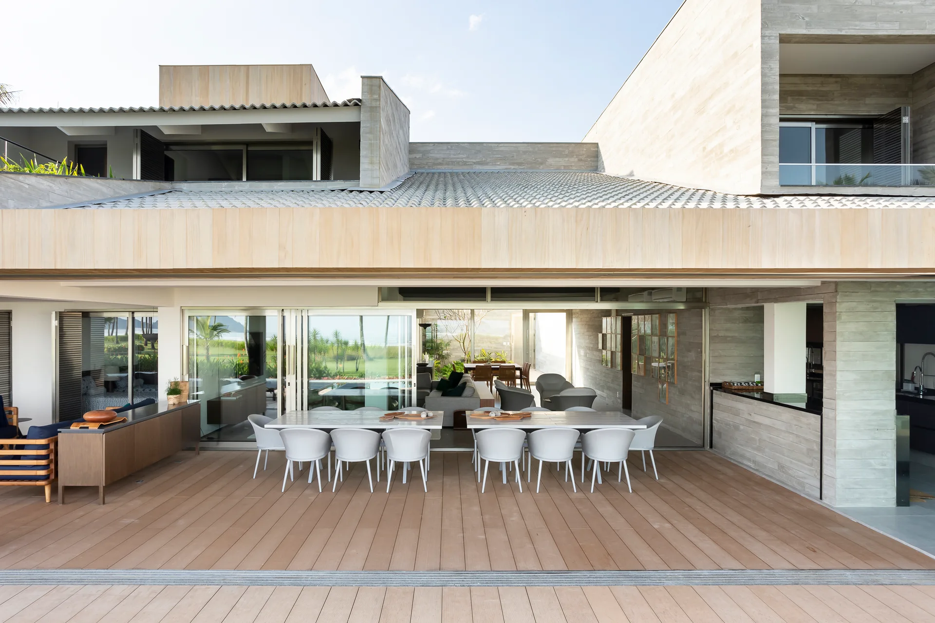 A modern outdoor patio features a wooden deck with a long white dining table and chairs beneath a covered roof overhang next to open sliding glass doors.