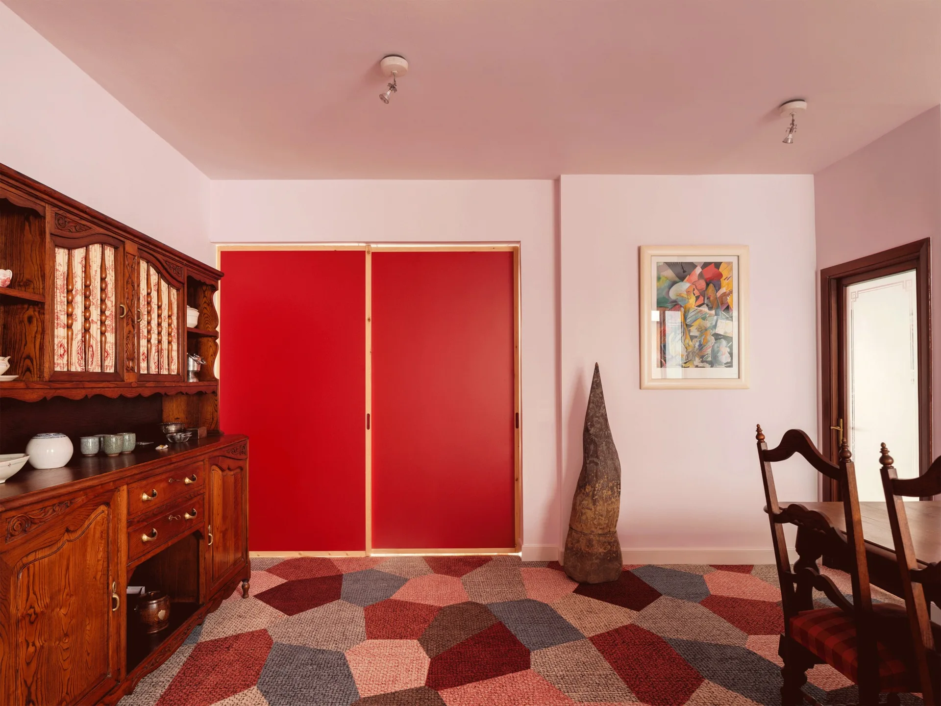 An eclectic dining room features pale pink walls, a large red sliding door, a vintage wood hutch, and a bold geometric patterned carpet.