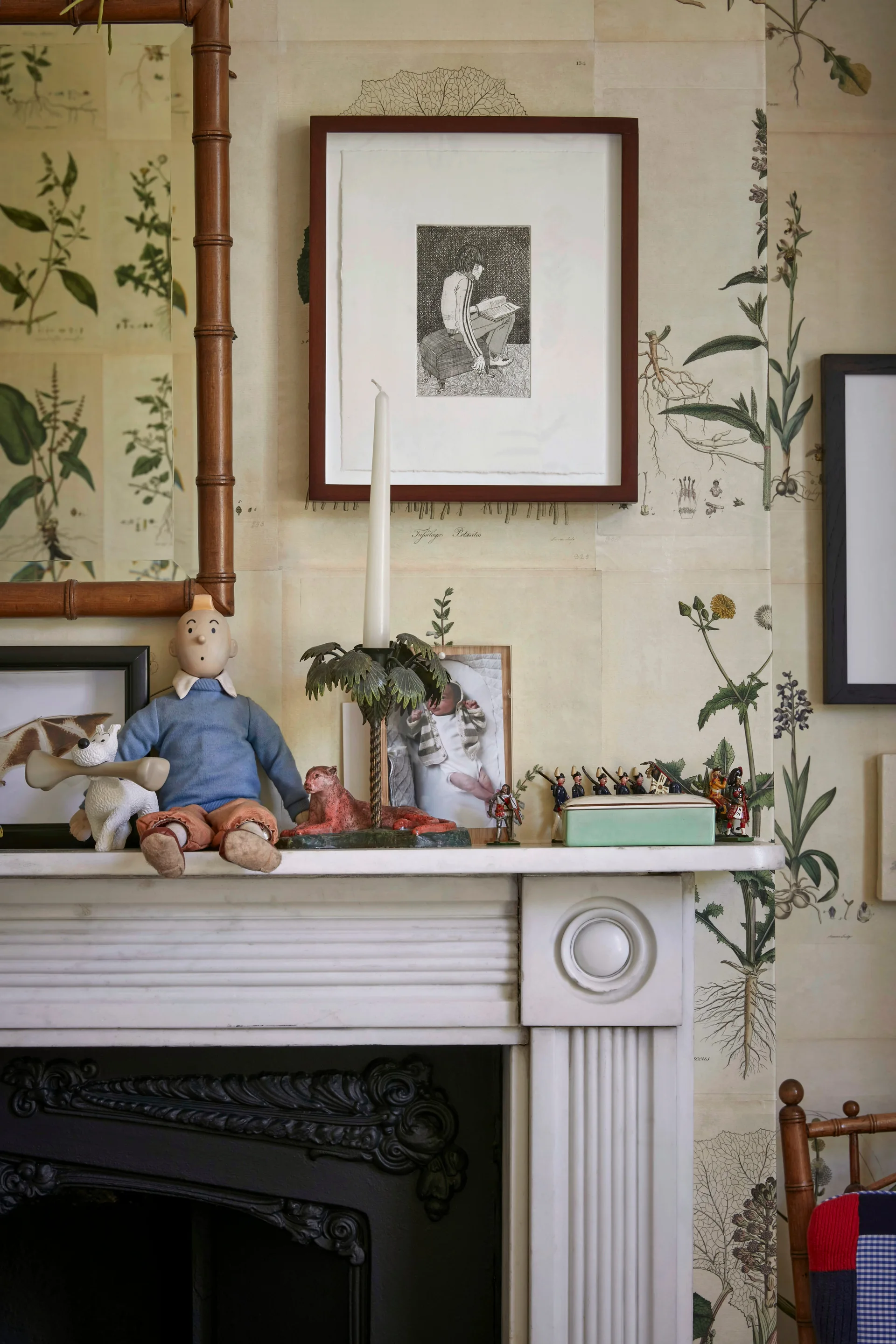 A painted white mantelpiece displays diverse vintage figurines and a framed drawing against cream botanical illustration wallpaper.