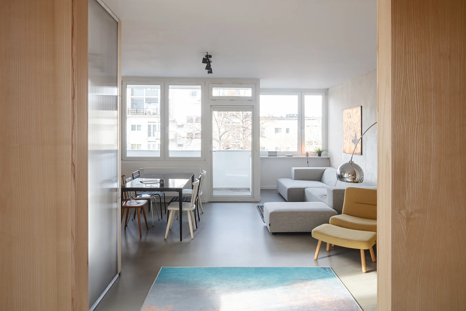An open-plan living and dining room with a grey sectional, a yellow armchair with ottoman, a glass dining table, and large back windows.
