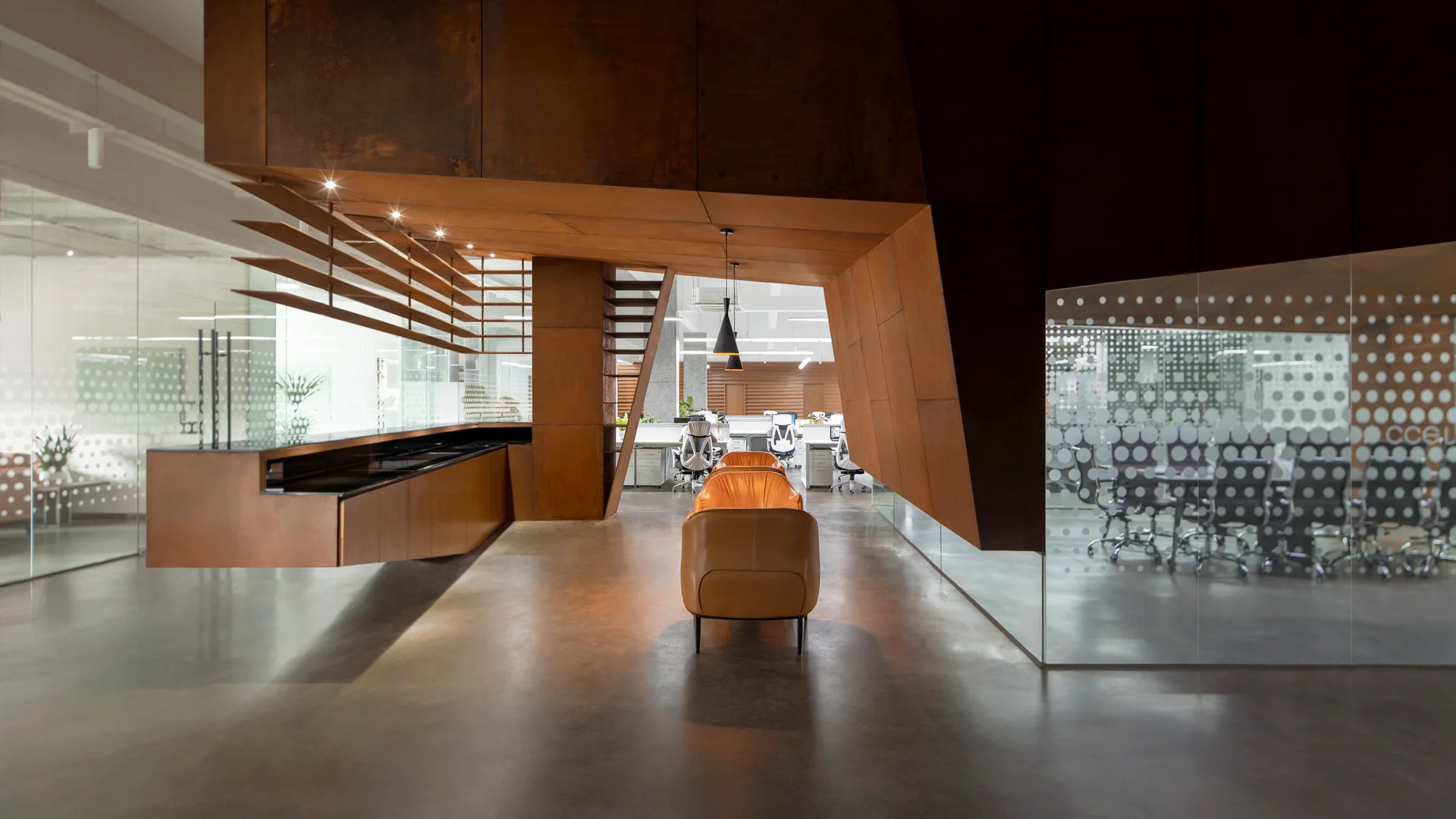 A modern open-plan office features a large weathered steel architectural structure, polished concrete floors, glass partitions, and tan leather seating.