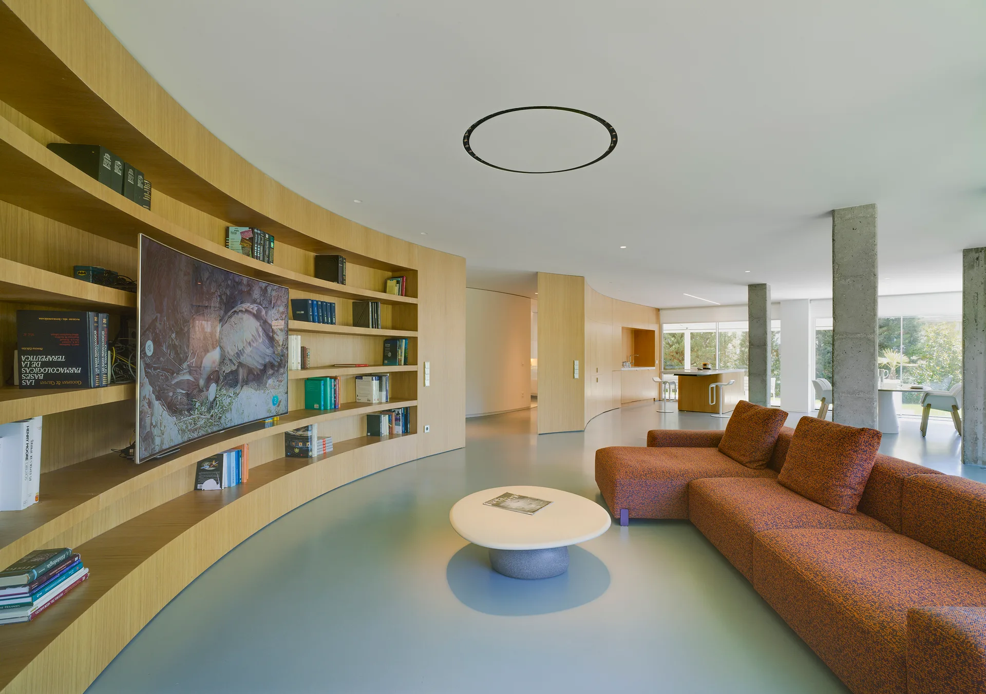 A modern open-plan living room featuring a long curved wood shelving wall with a television, a large orange sectional sofa, and concrete columns.
