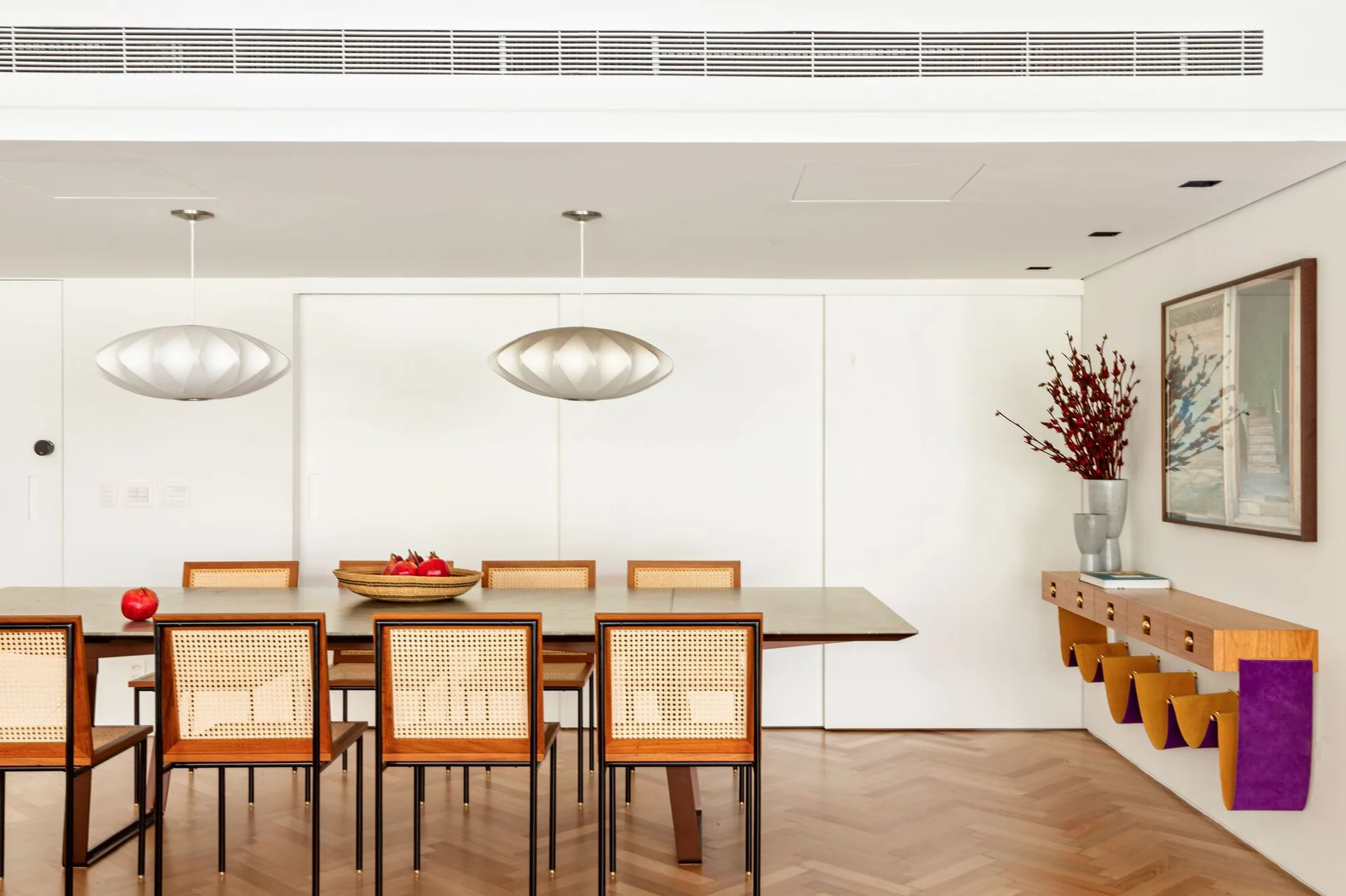 A modern dining room features a long table with cane-back chairs, herringbone wood flooring, and two elliptical pendant lights hanging overhead.