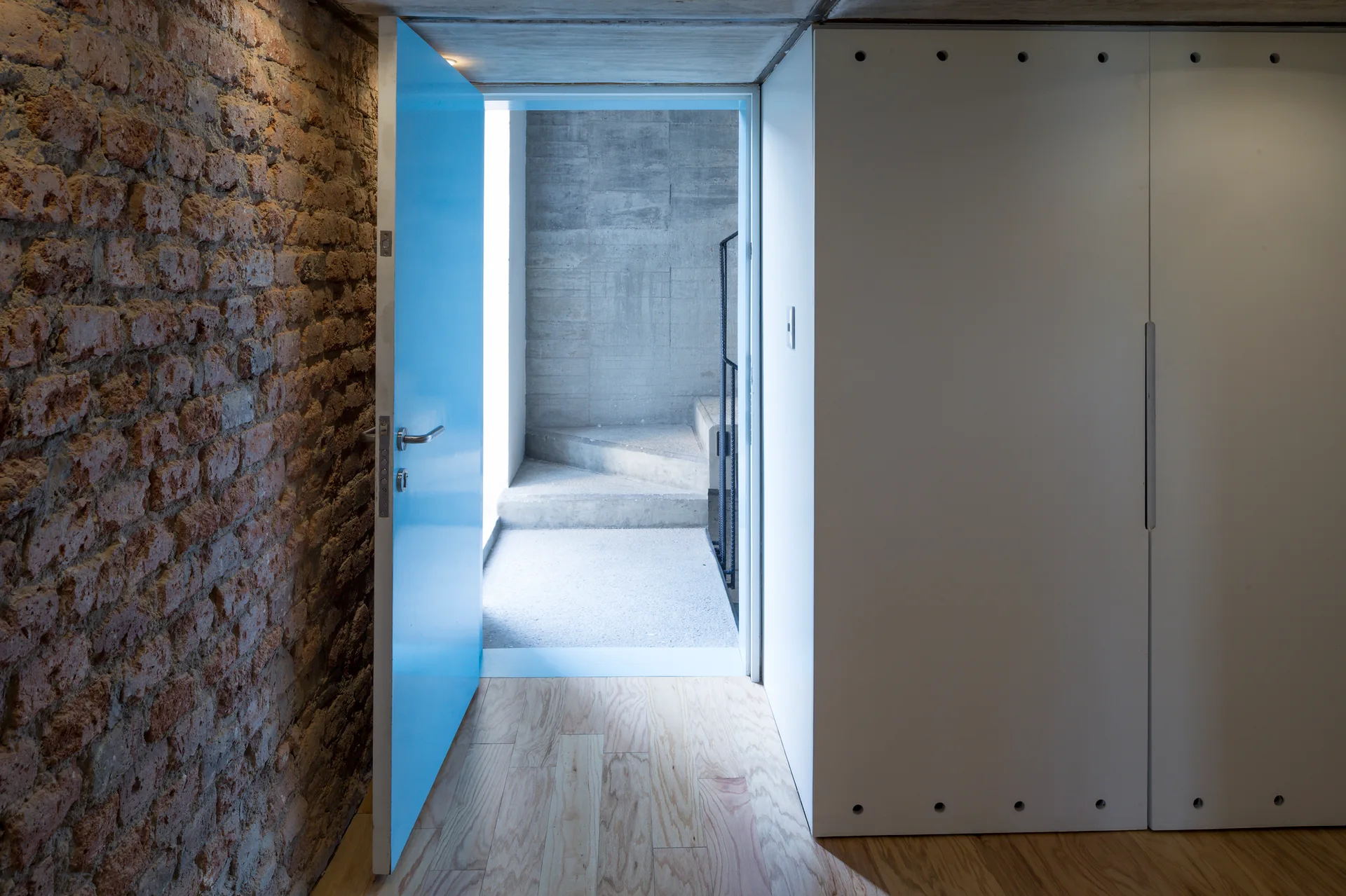 An industrial entryway features an exposed brick wall, a bright blue door, light wood floors, and a white closet system leading to concrete stairs.