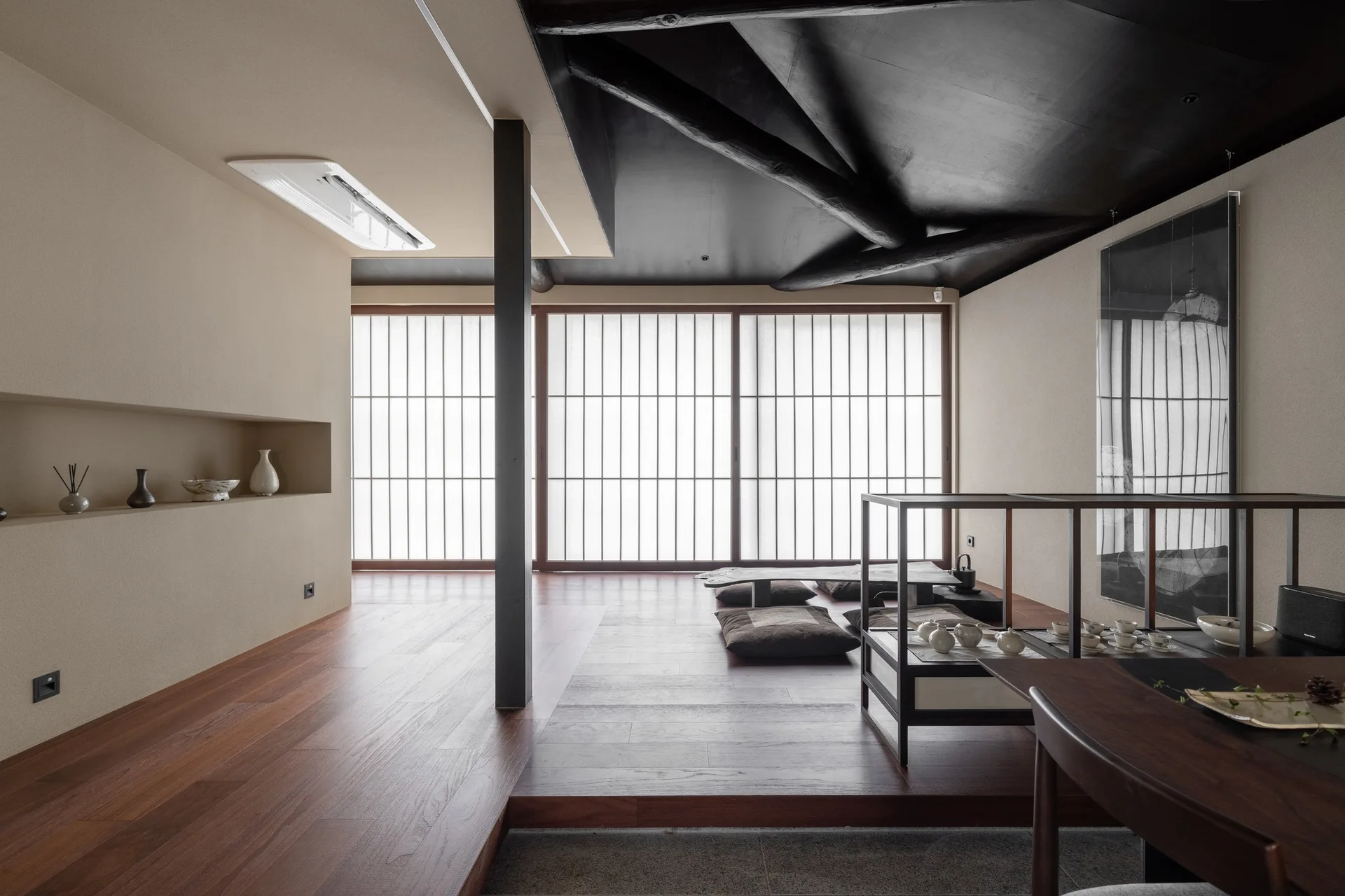 A minimalist Japanese style tea room with a raised wood floor platform, floor cushions, sliding shoji screens, and a dark beamed ceiling.