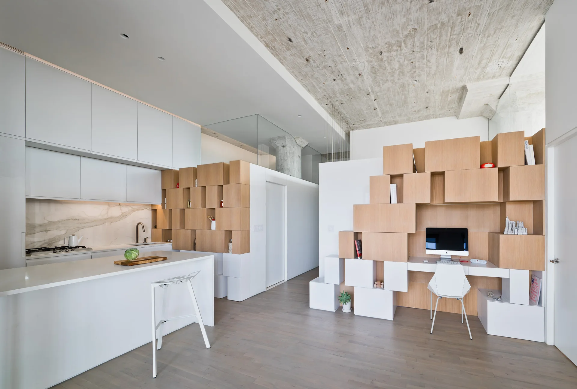A modern open-plan interior featuring a sleek white kitchen island next to an integrated wooden modular desk unit under an exposed concrete ceiling.