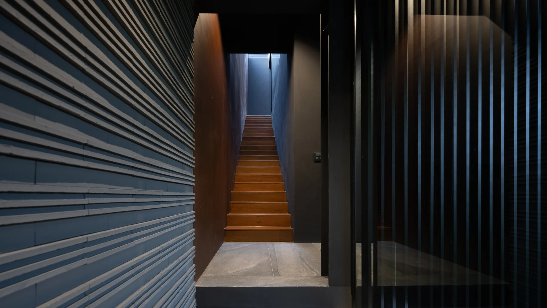 A dark modern staircase with warm wood treads rises between a textured horizontal paneled wall and a black vertical slatted screen.