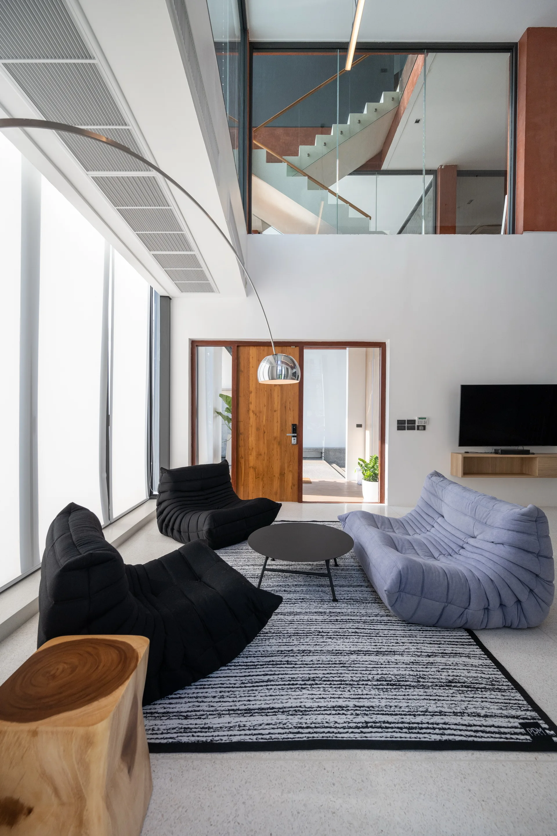 A modern living room featuring low-slung black and blue modular sofas on a striped rug with an arched floor lamp and staircase visible upstairs.