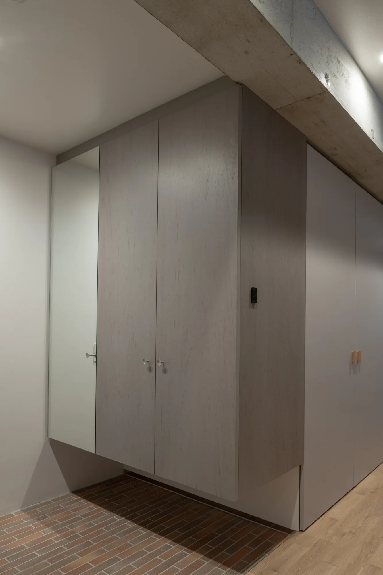 A minimalist entryway features a light wood wardrobe with a built-in mirror, an exposed concrete ceiling beam, and brick tile flooring.