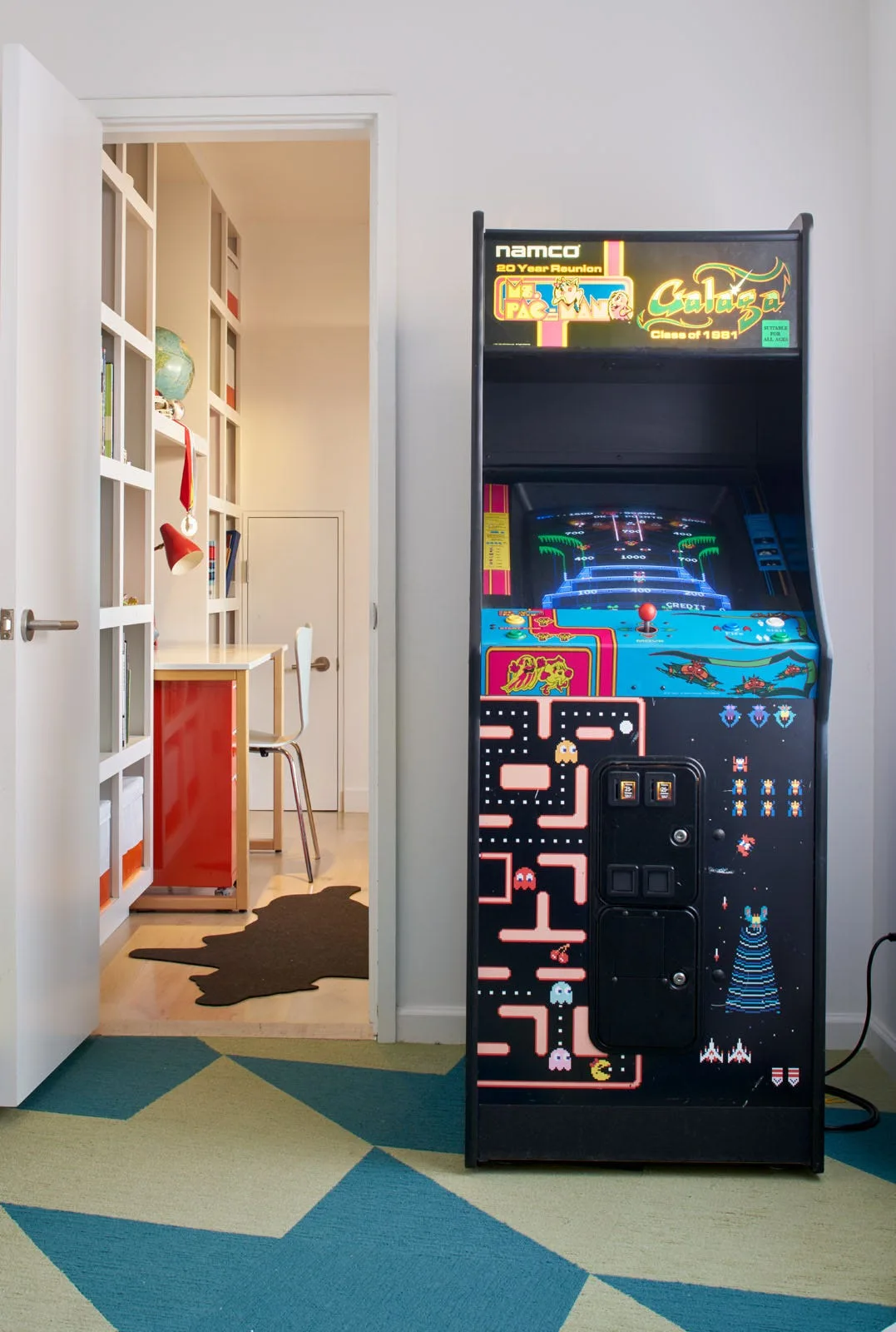 A retro arcade cabinet stands on a bold geometric carpet next to an open doorway showing a study room with built-in bookshelves.