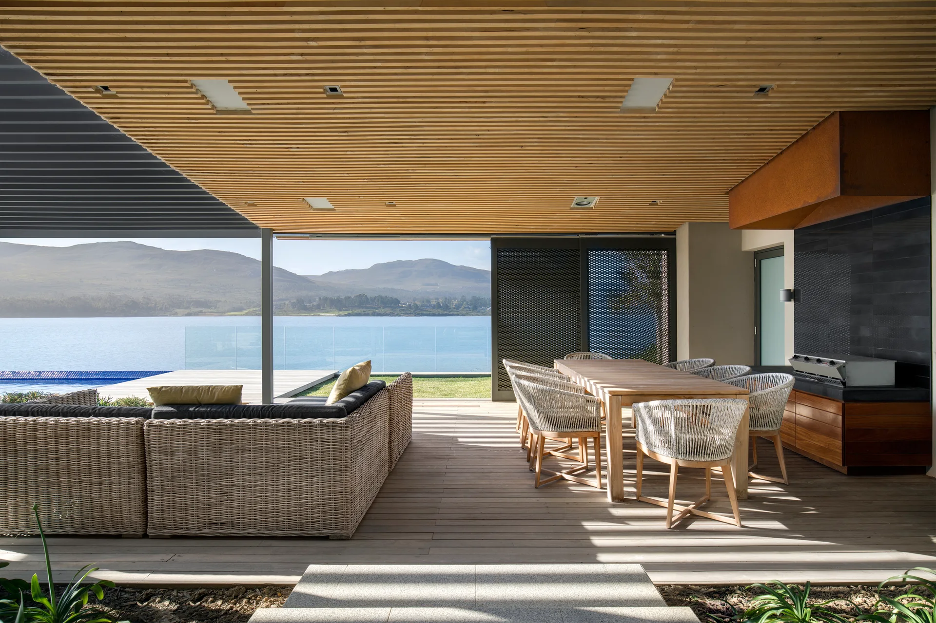 A covered waterfront patio features a wood slatted ceiling, a wooden dining table with woven chairs, a wicker sectional, and an outdoor kitchen area.