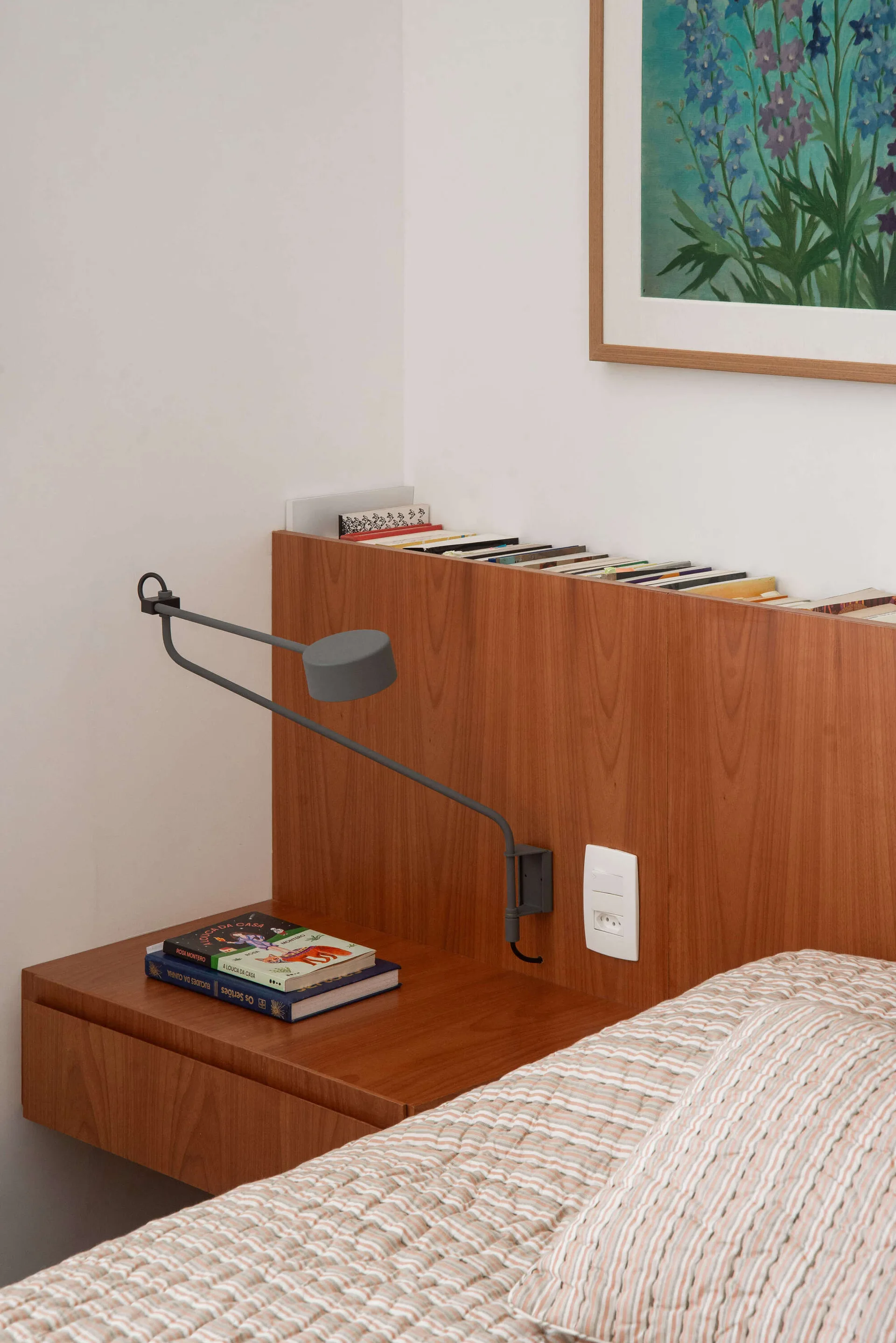 A close-up of a wooden headboard in a bedroom featuring integrated book storage on top, a grey articulating lamp, and a floating nightstand drawer.