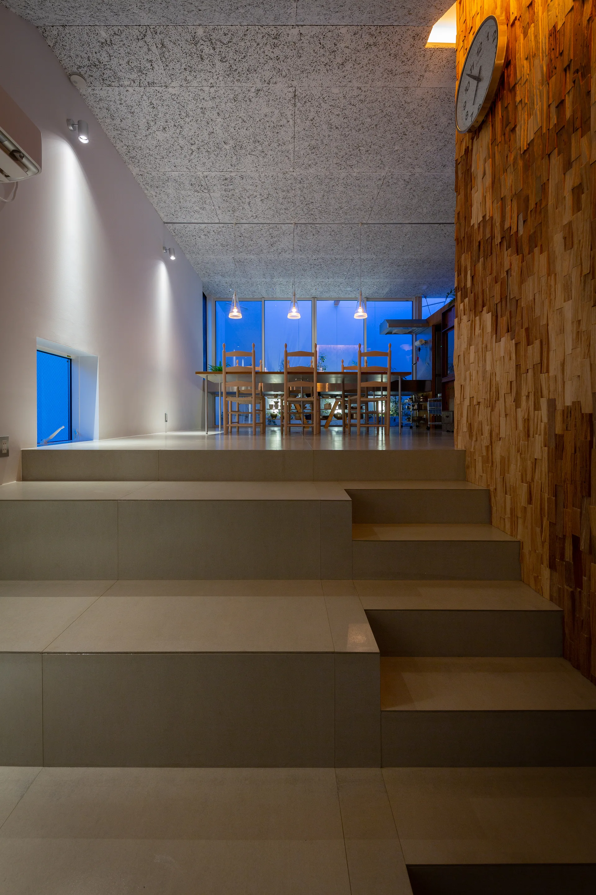 Broad beige tile steps lead up to an elevated dining room featuring a textured wood accent wall, three pendant lights, and large windows at twilight.