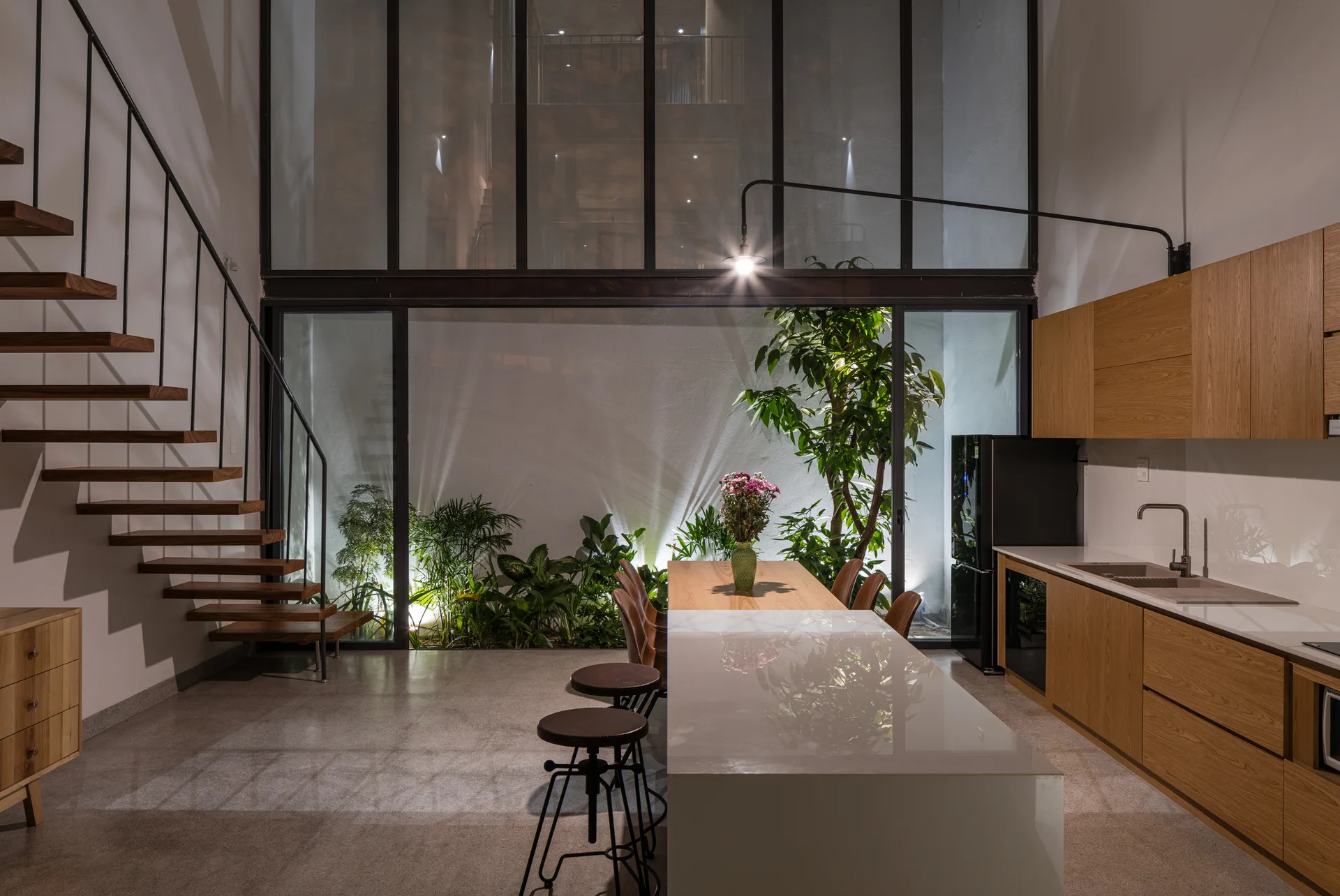 A modern kitchen features a glossy white island, wooden cabinets, a floating wooden staircase, and an indoor garden visible through a large glass wall.