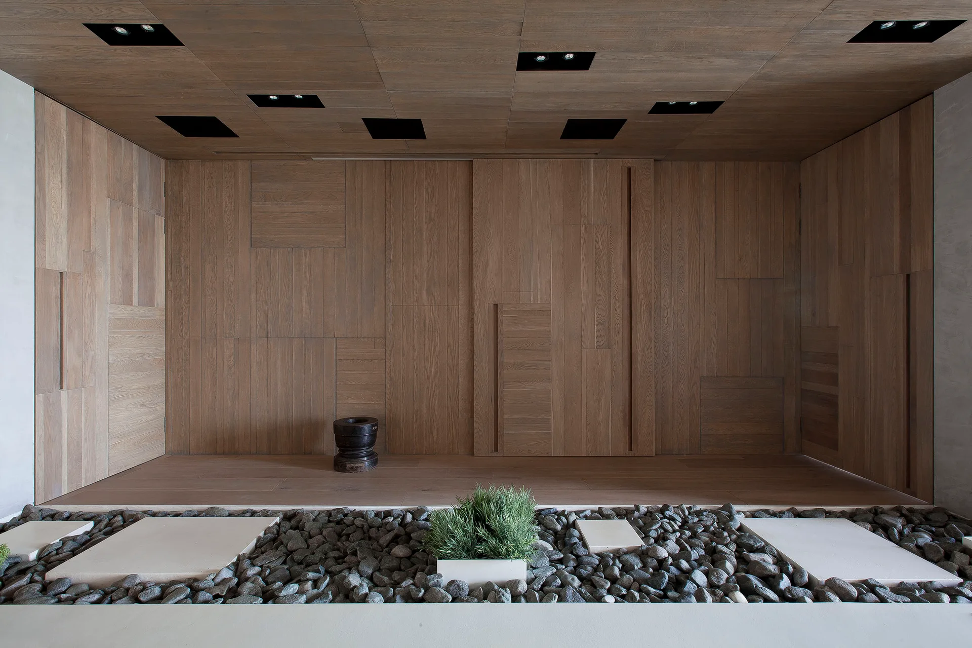 A minimalist wood-paneled room features an indoor pebble garden with a small plant, stepping stones, and recessed square ceiling lights.