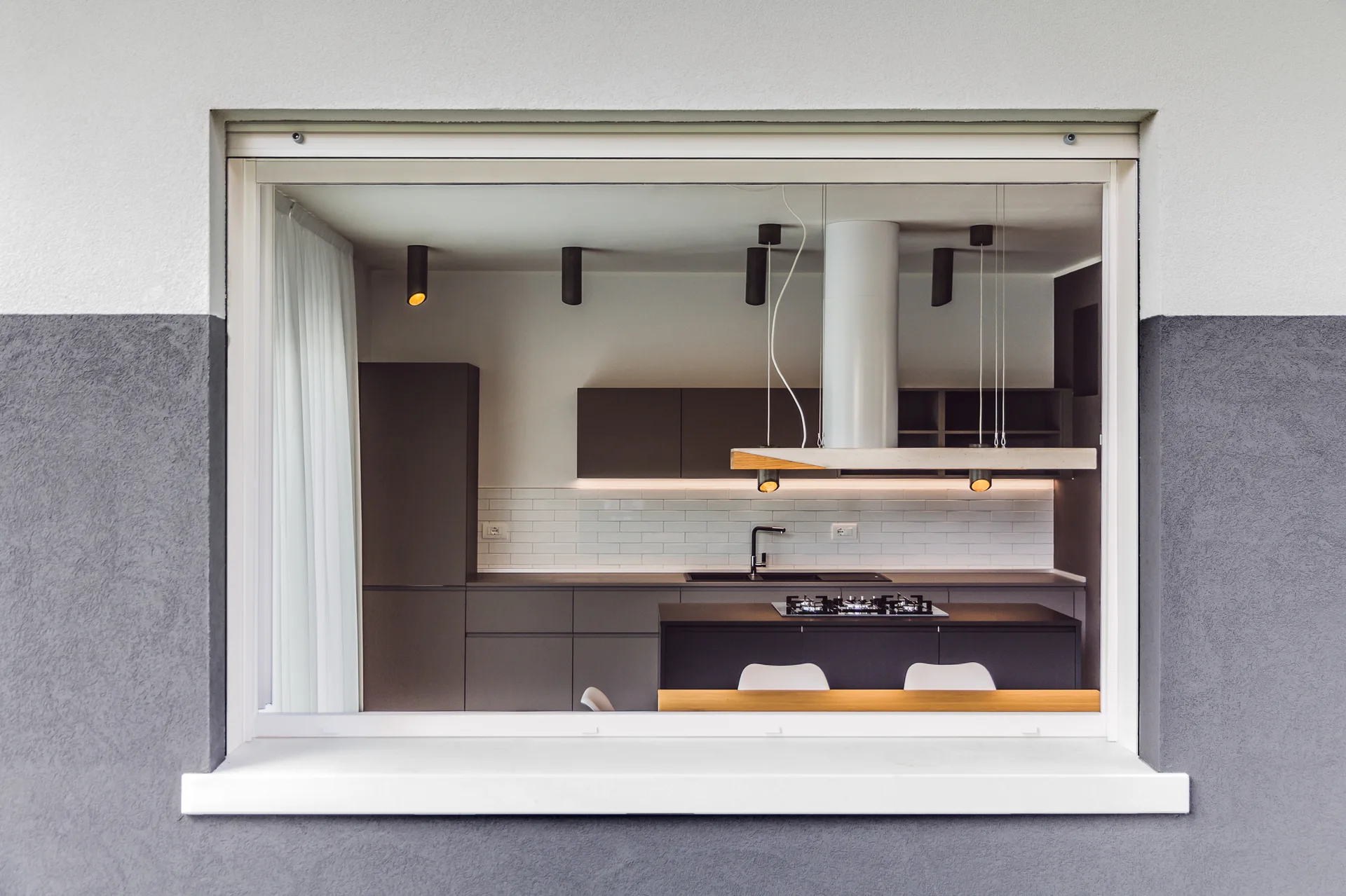 A modern kitchen with matte grey cabinetry and a white subway tile backsplash, viewed through a large white-framed window.