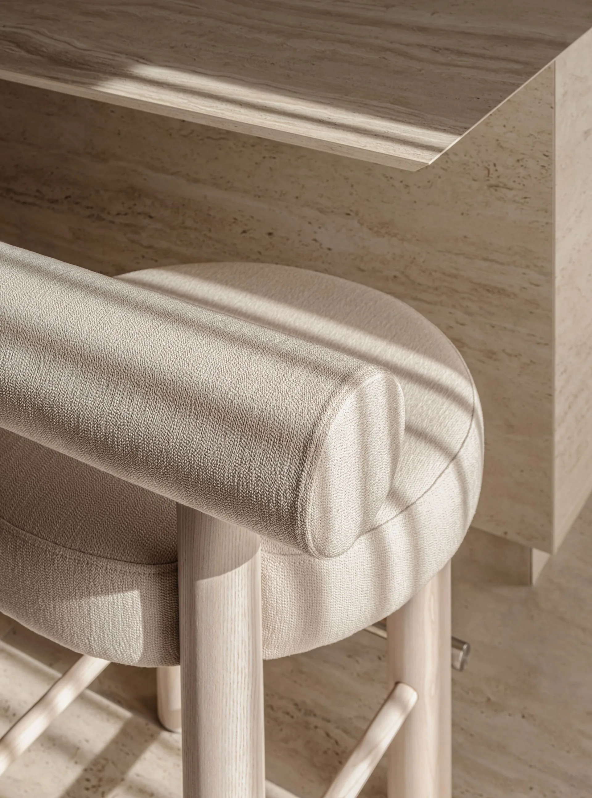 A close-up of a cream upholstered stool with a cylindrical bolster back and light wood legs, positioned next to a beige travertine stone desk.