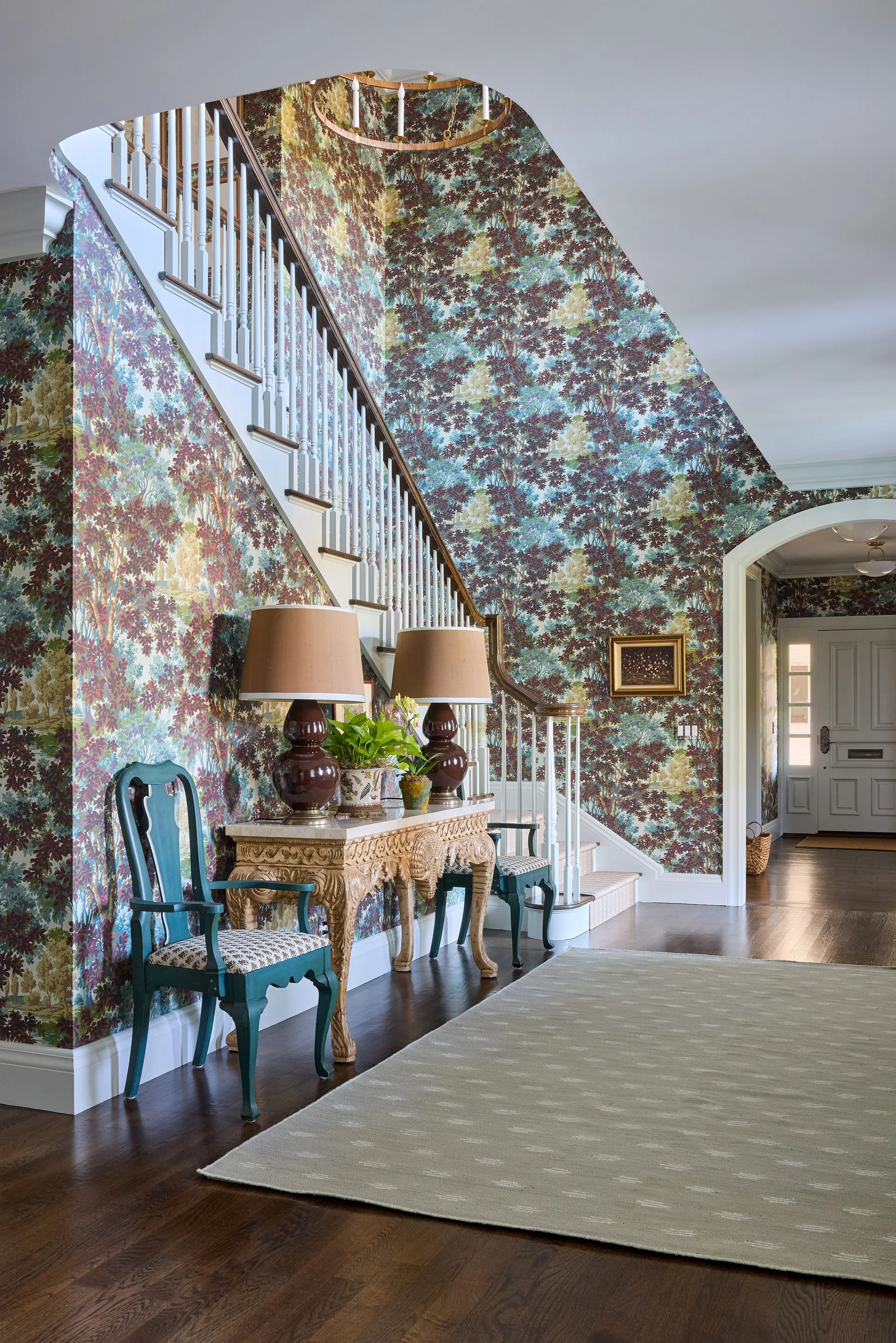 An entryway features walls covered in a green and brown tree pattern wallpaper, an ornate wood console table flanked by two teal chairs, and a staircase.