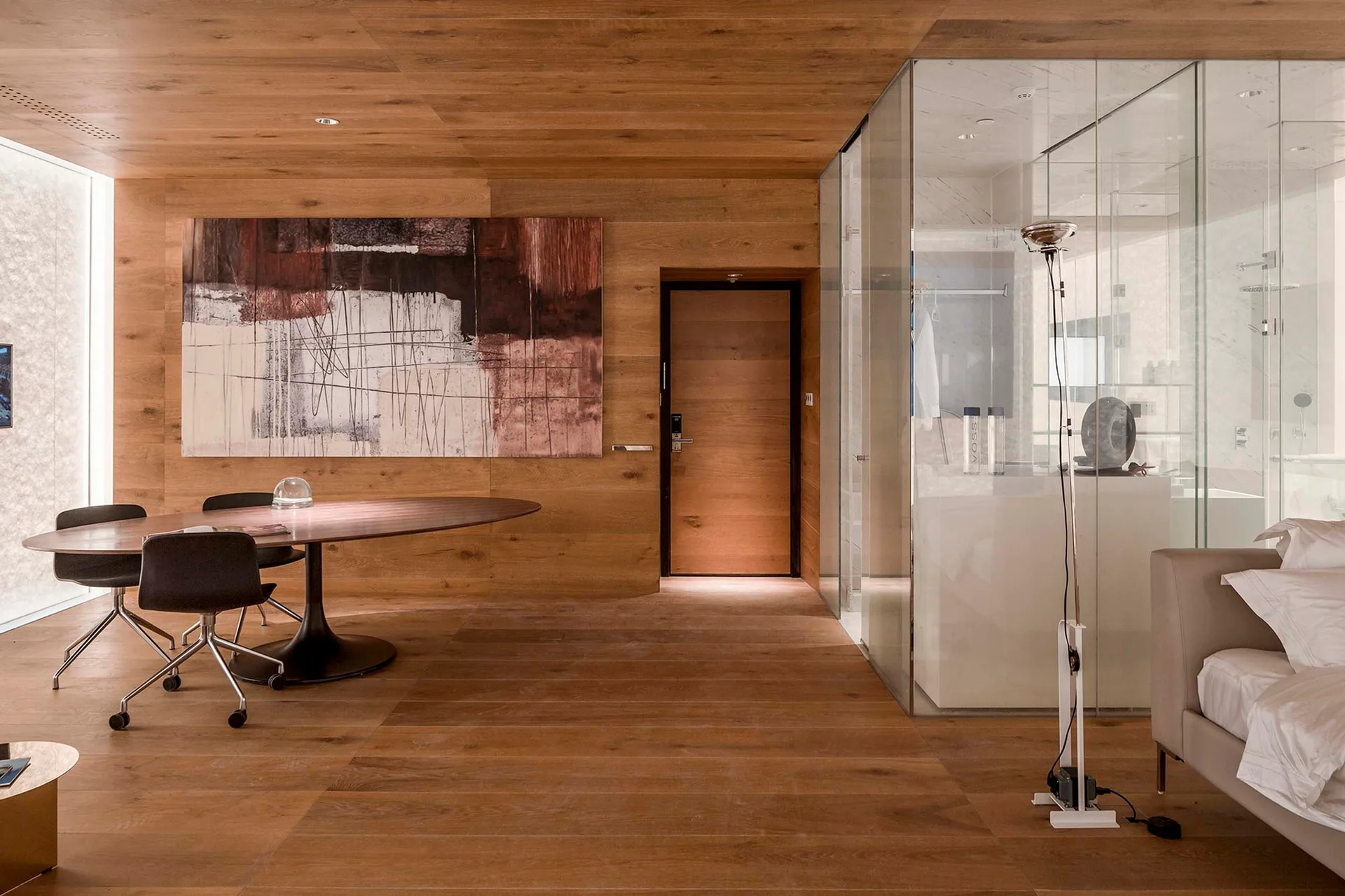 A modern bedroom suite with warm wood-paneled walls and ceiling, featuring an oval dining table and a glass-enclosed bathroom partition.