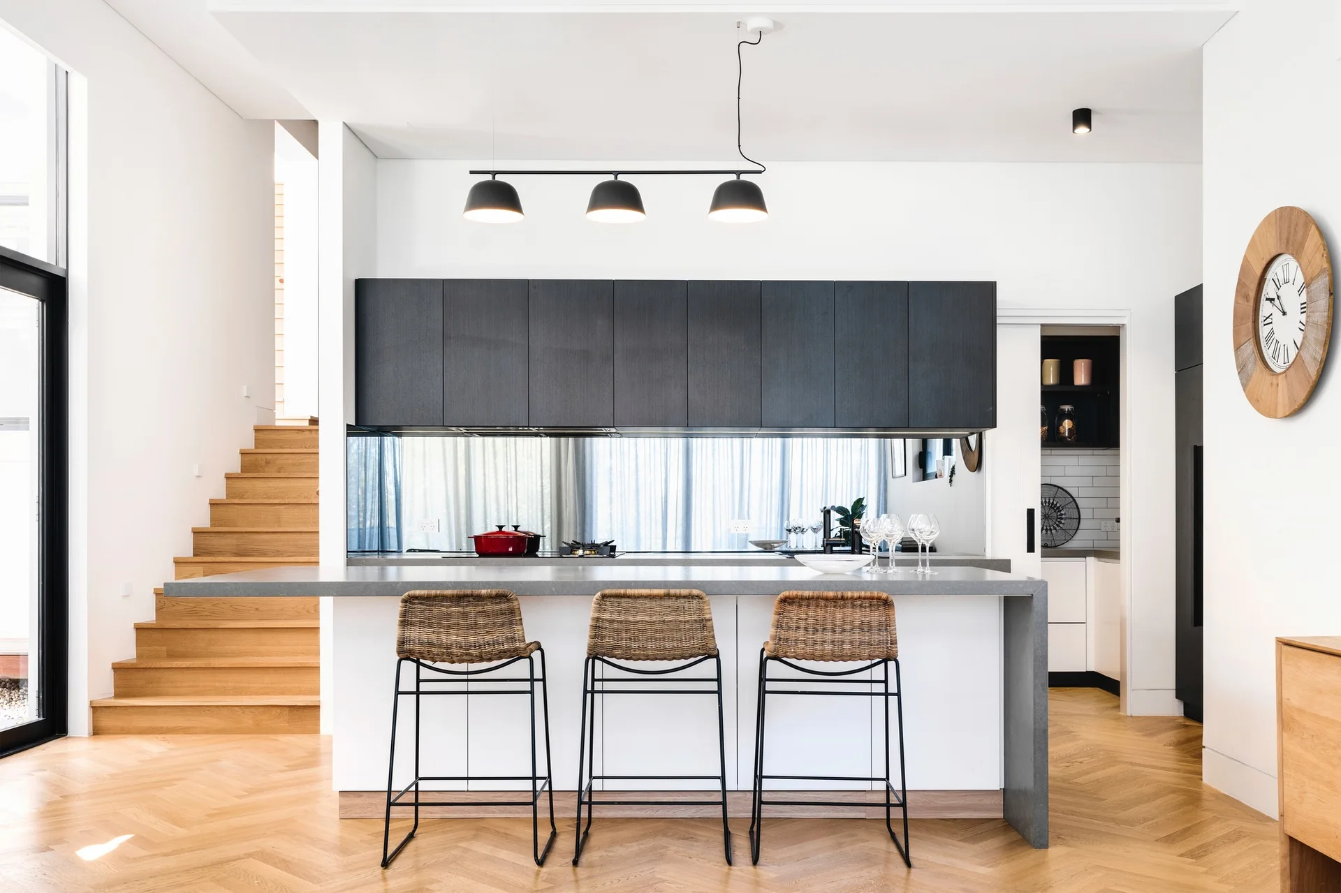 A modern kitchen features a white island with woven stools, dark upper cabinets, a mirrored backsplash, and herringbone wood flooring.