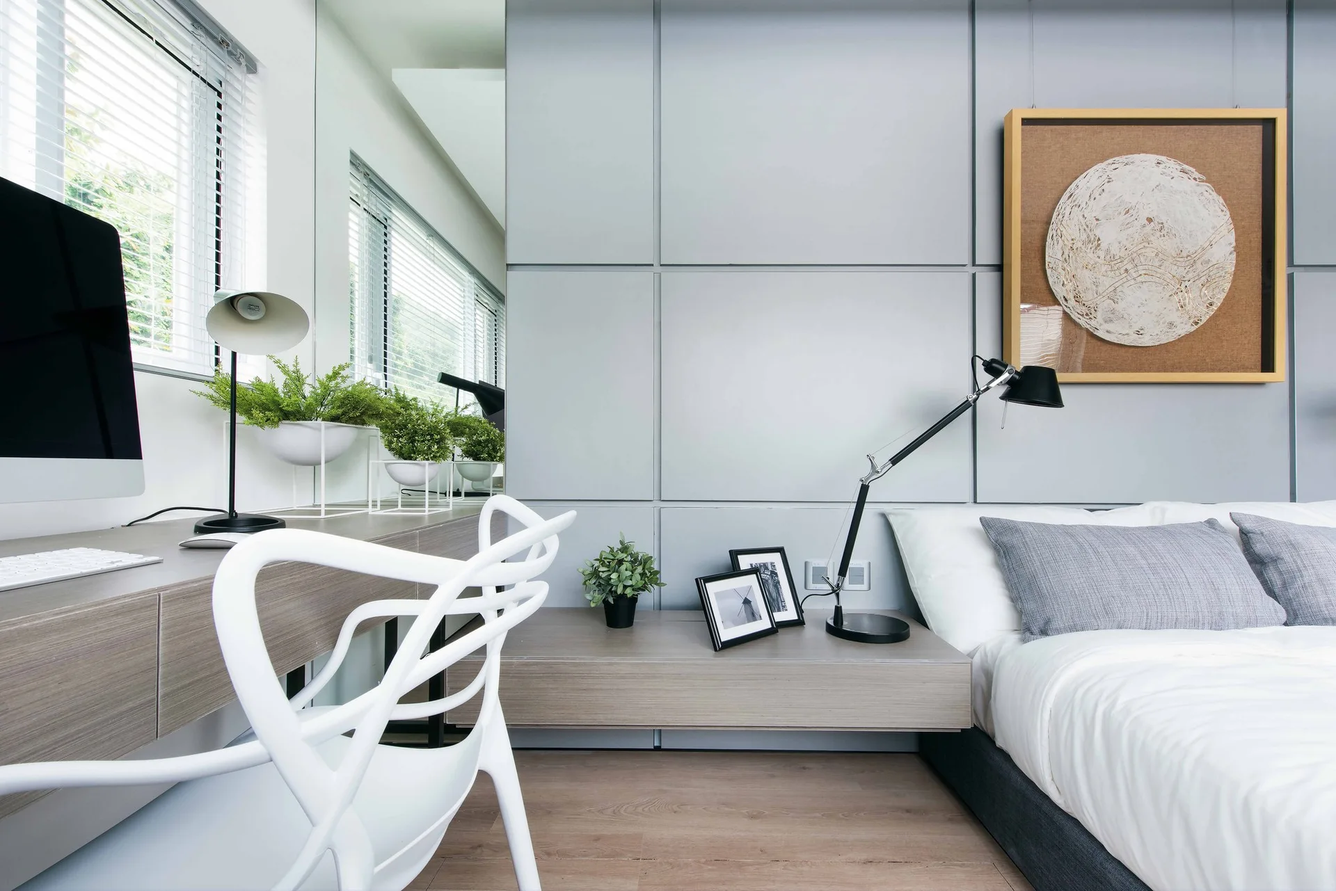 A modern bedroom featuring a grey grid paneled wall, a bed with white bedding, and an integrated wood desk with a white organic chair.