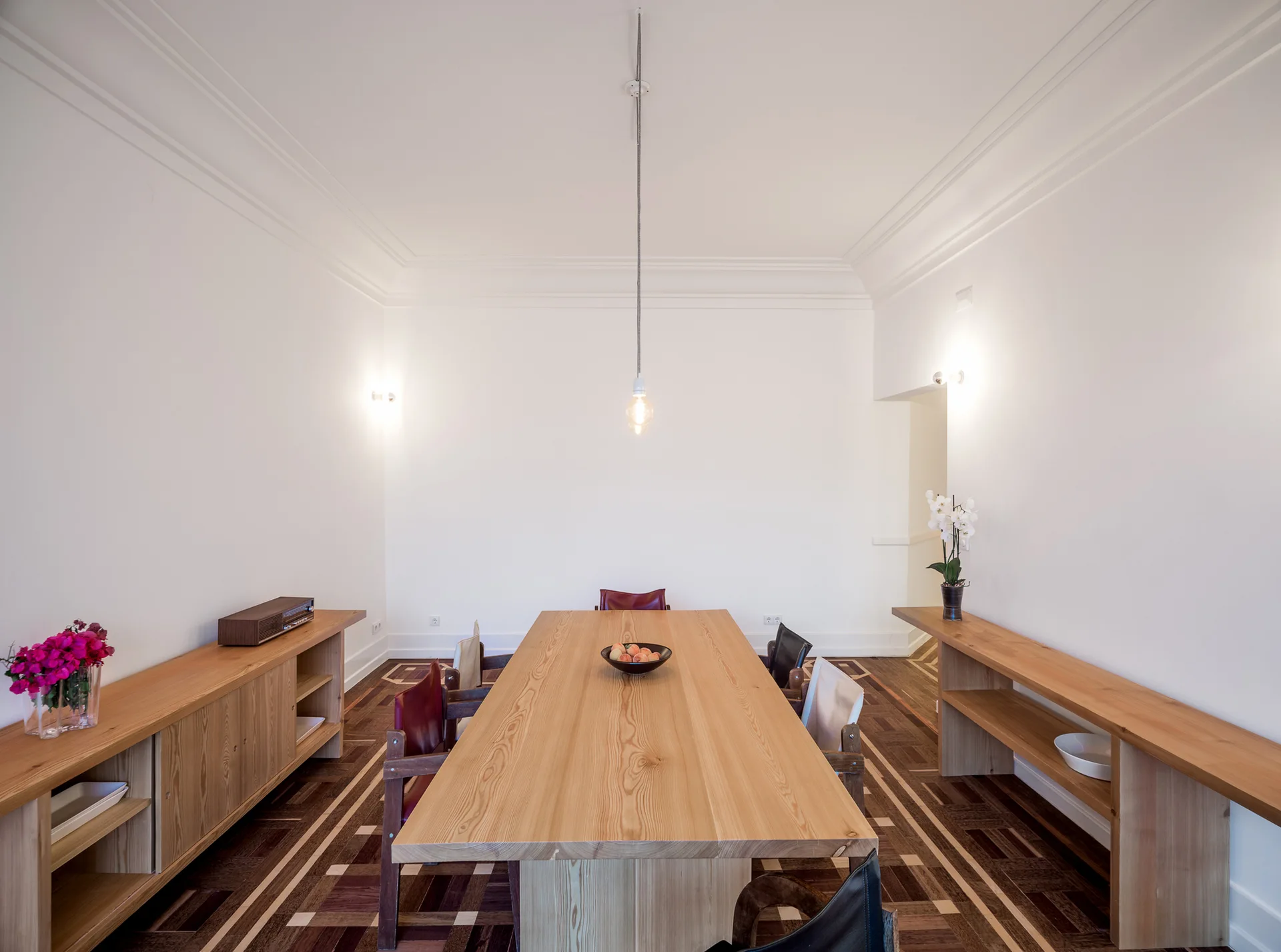 A minimalist dining room features a long light wood table, matching side consoles, and a geometric pattern parquet wood floor.