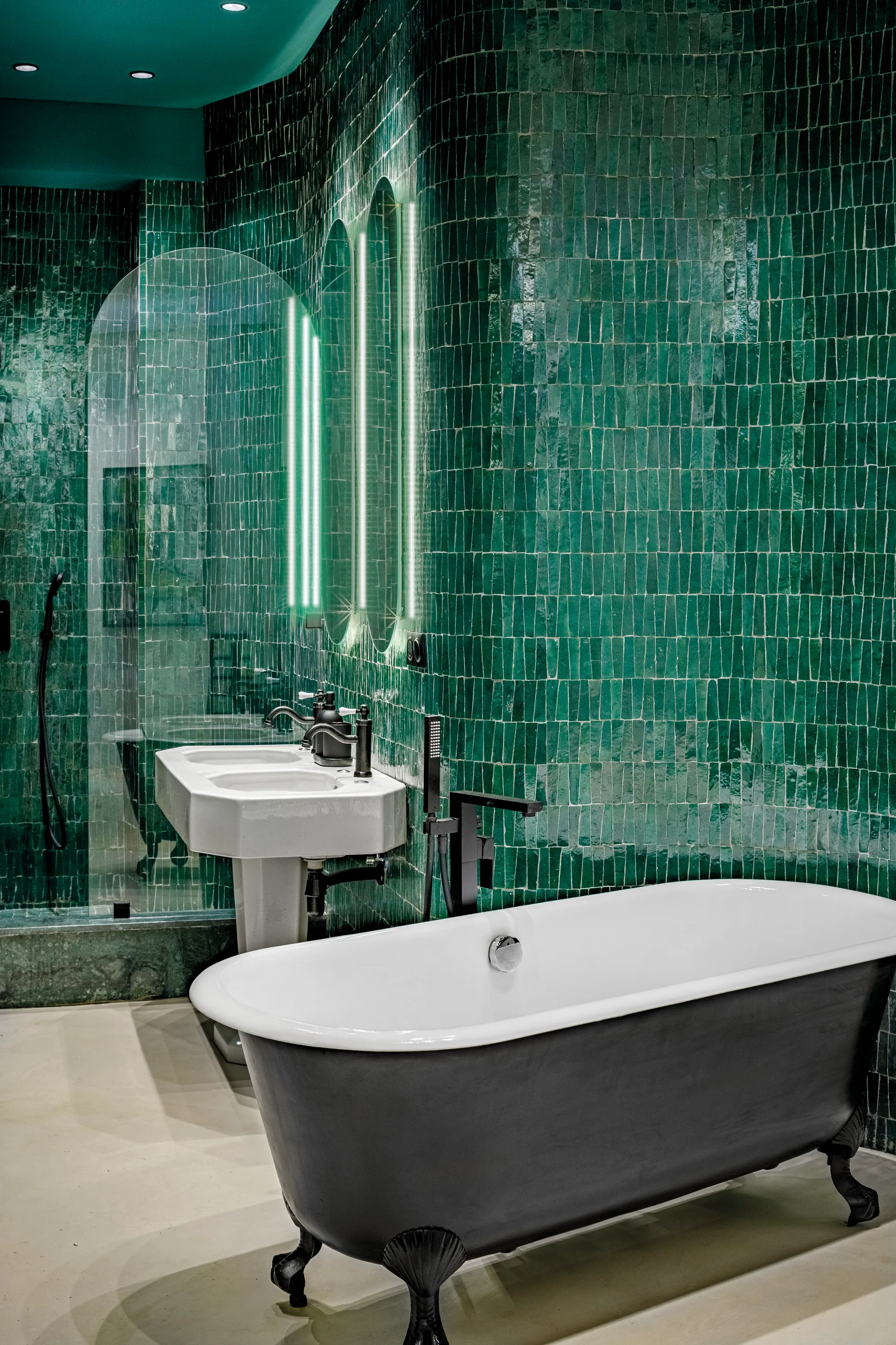 A bathroom with glossy emerald green tiled walls, a black and white clawfoot bathtub, and a pedestal sink under arched backlit mirrors.