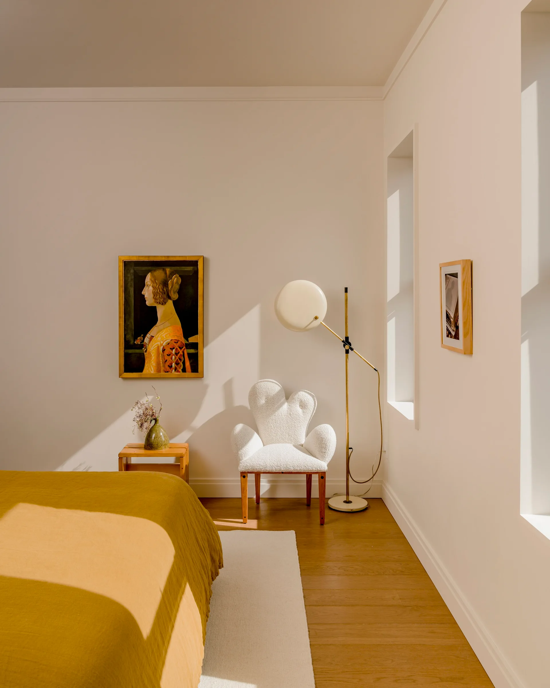 A sunlit bedroom with a mustard yellow bed, a textured white boucle armchair, a brass floor lamp, and a framed portrait on the wall.