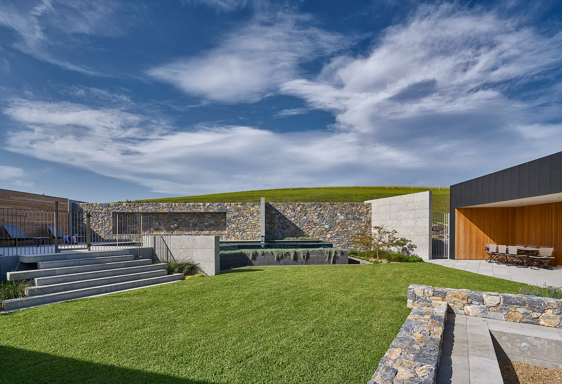 A modern backyard landscape features a manicured green lawn, a textured stone feature wall, a swimming pool, and a covered wood-clad patio.
