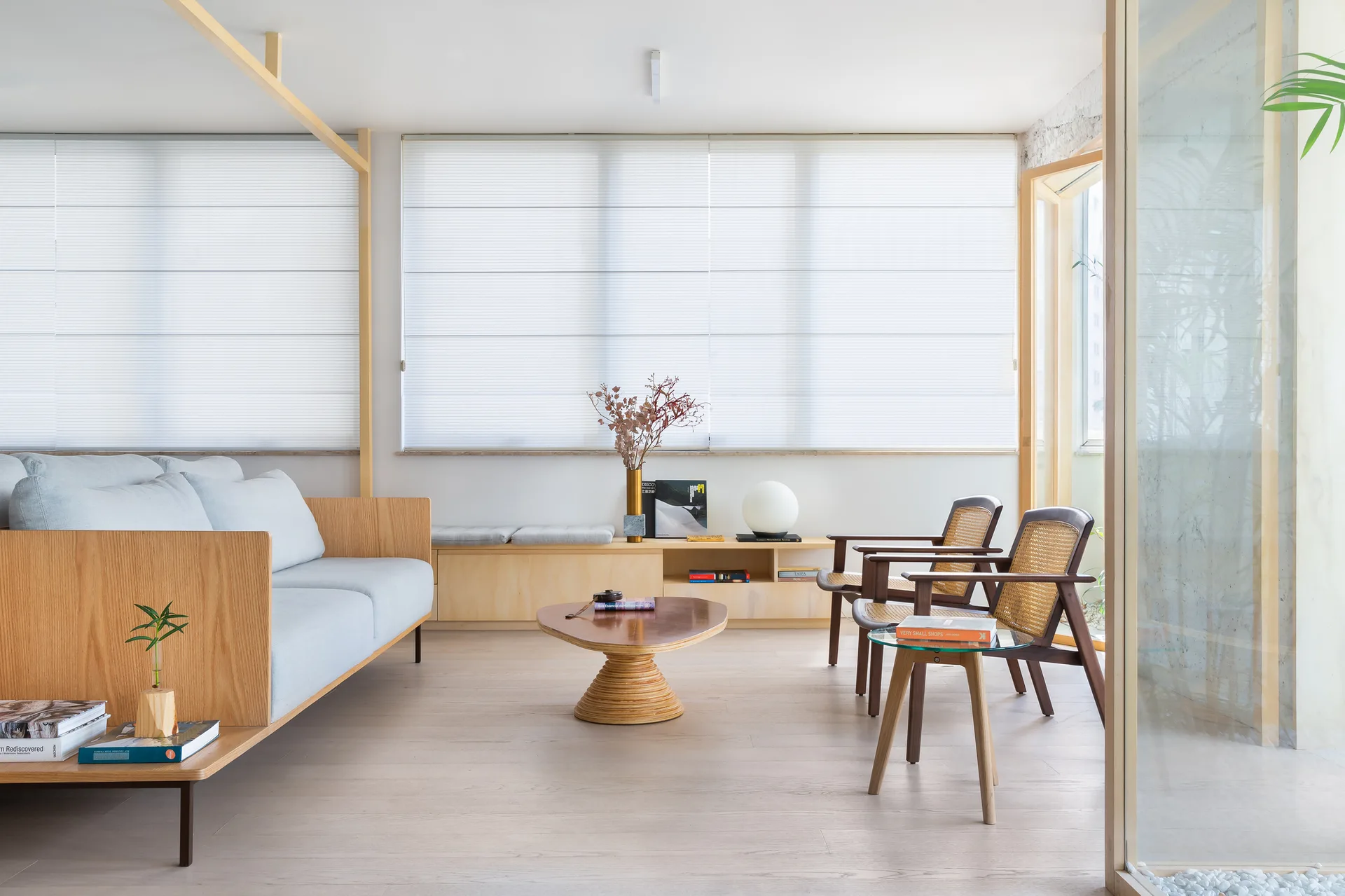 A light-filled minimalist living room features a wood-framed grey sofa, an organic wooden coffee table, and two cane-back armchairs.