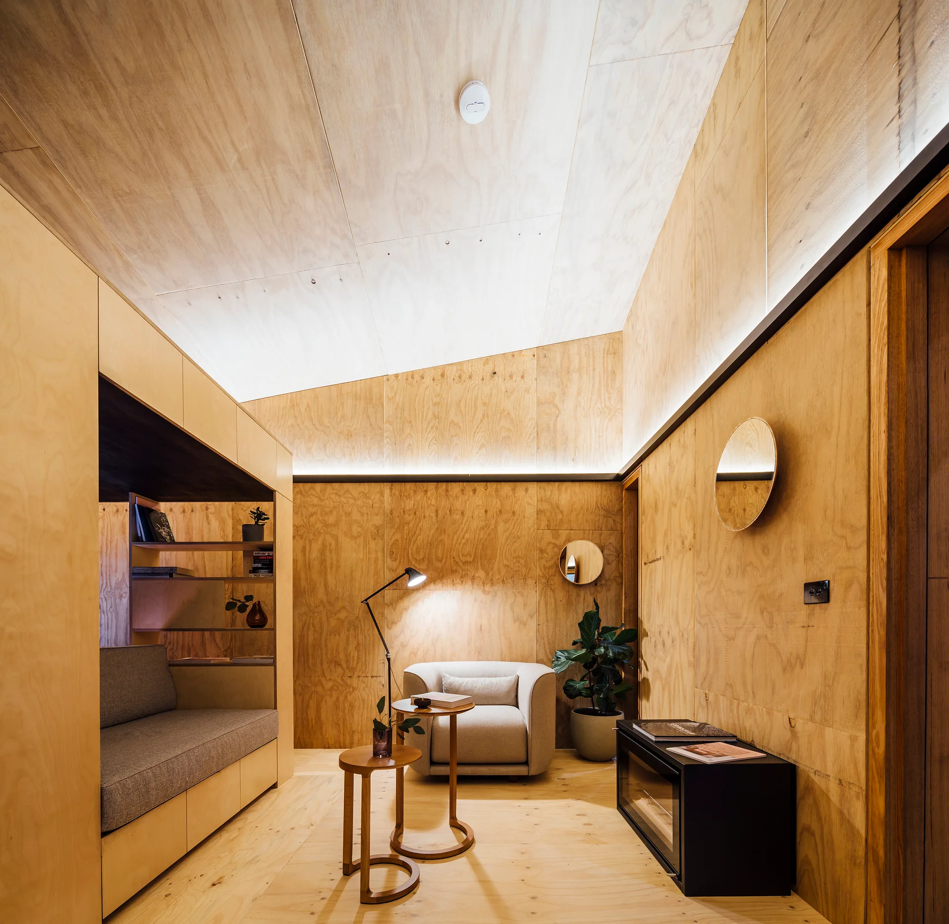 A minimalist bedroom featuring walls and an angled ceiling clad in warm plywood paneling, with a built-in sofa nook and an armchair.