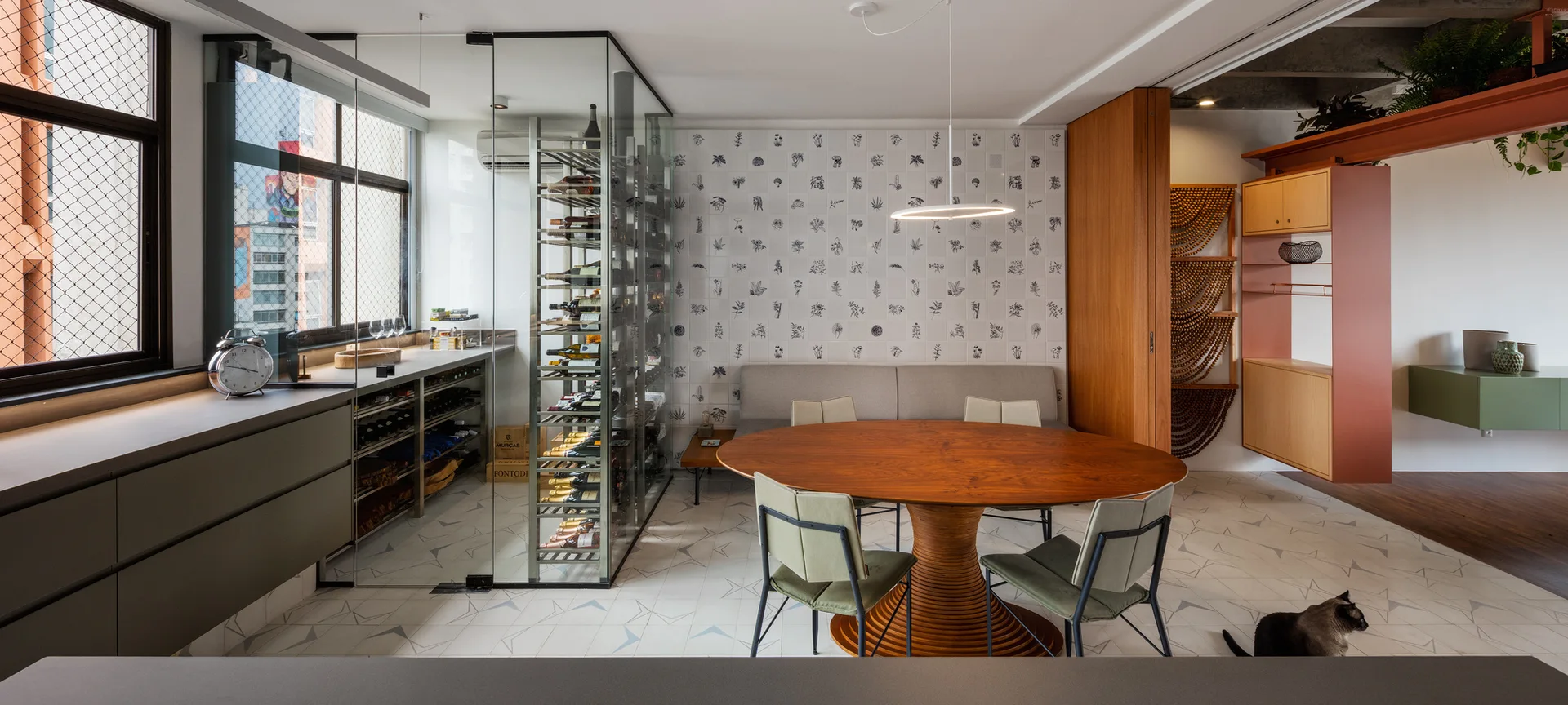 A contemporary dining room featuring a round wood pedestal table, green chairs, and a glass-enclosed wine storage unit next to a patterned wall.