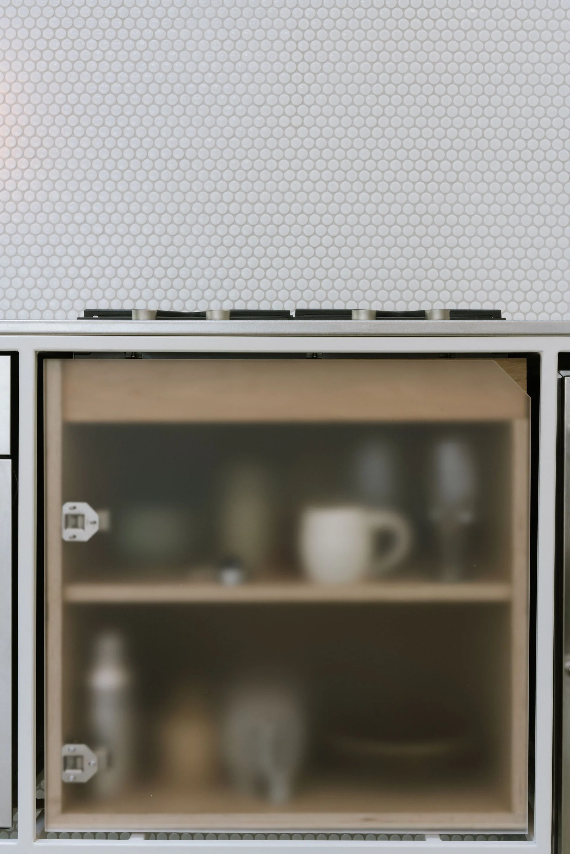 A minimalist kitchen cabinet with a frosted glass door showing wooden interior shelves, positioned beneath a white penny tile backsplash.