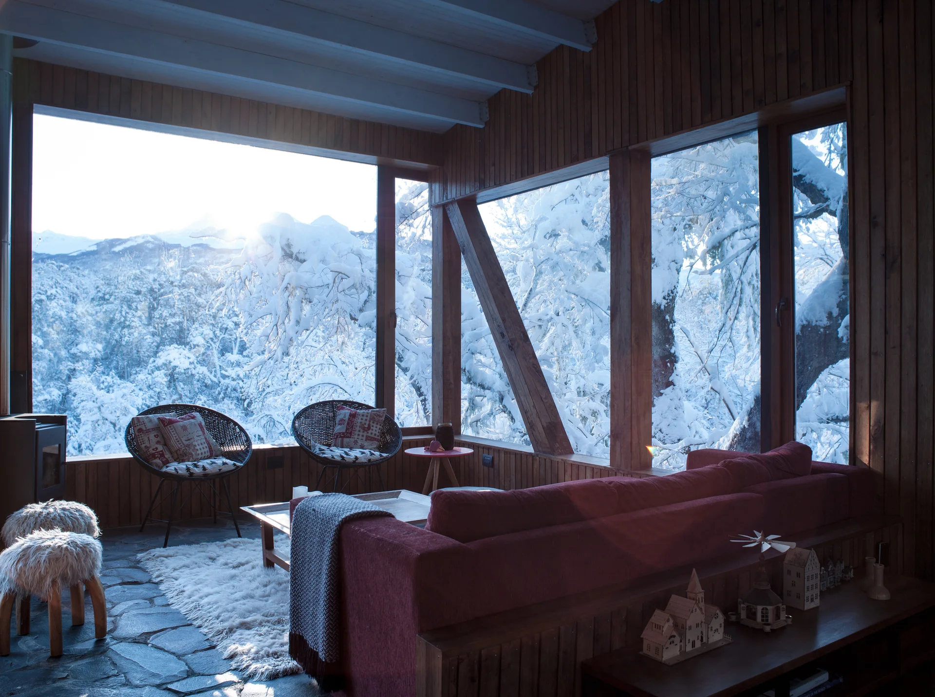 A rustic chalet living room features a red sofa and woven chairs on flagstone floors before large windows framing a snowy forest.