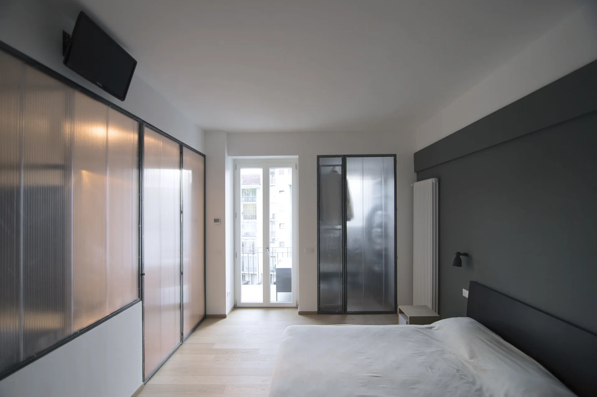 A modern bedroom with light wood floors, a dark grey accent wall behind the bed, and tall wardrobes with fluted glass sliding doors.